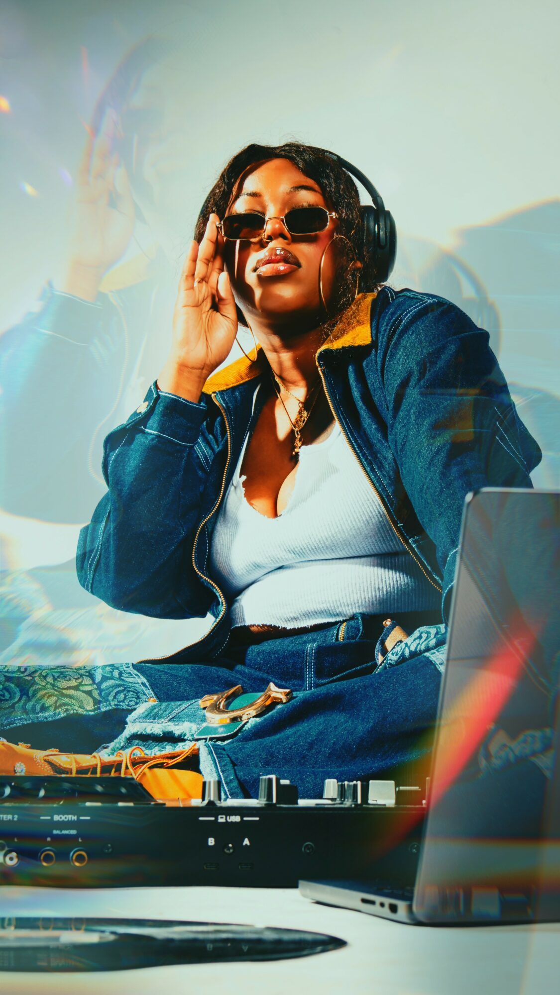 Woman DJ wearing sunglasses and headphones, smiling, with DJ equipment and laptop in front.