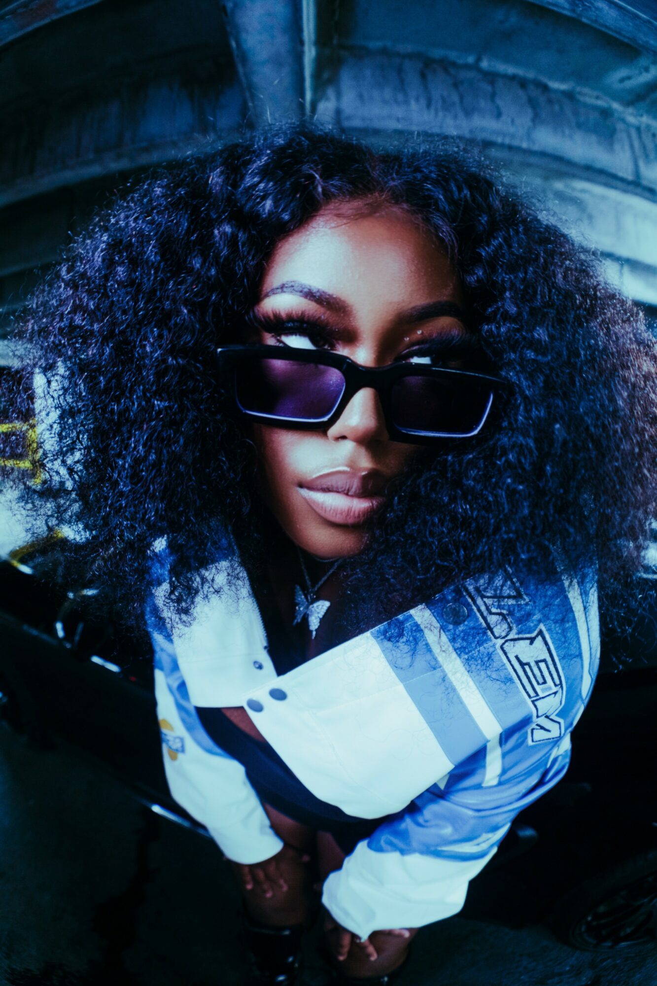 Person with curly hair wearing sunglasses and a white and blue jacket, looking up at the camera.