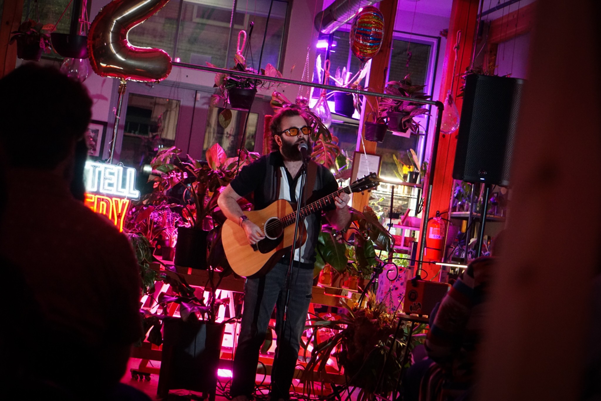 Man playing guitar on stage with pink and purple lighting, surrounded by plants and decorations, audience in foreground.