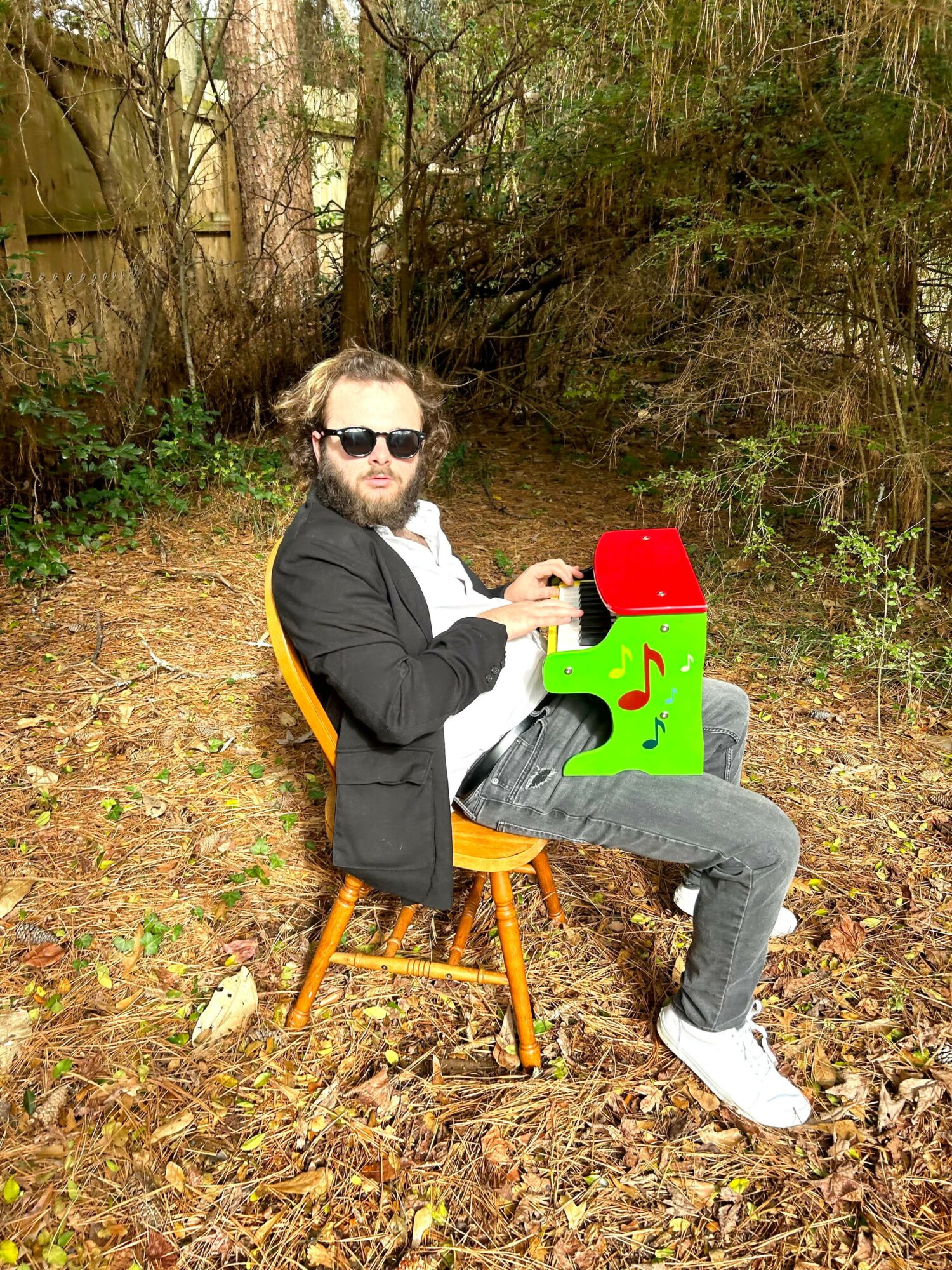 Man with sunglasses sitting on a wooden chair outdoors, playing a colorful toy, surrounded by trees and fallen leaves.