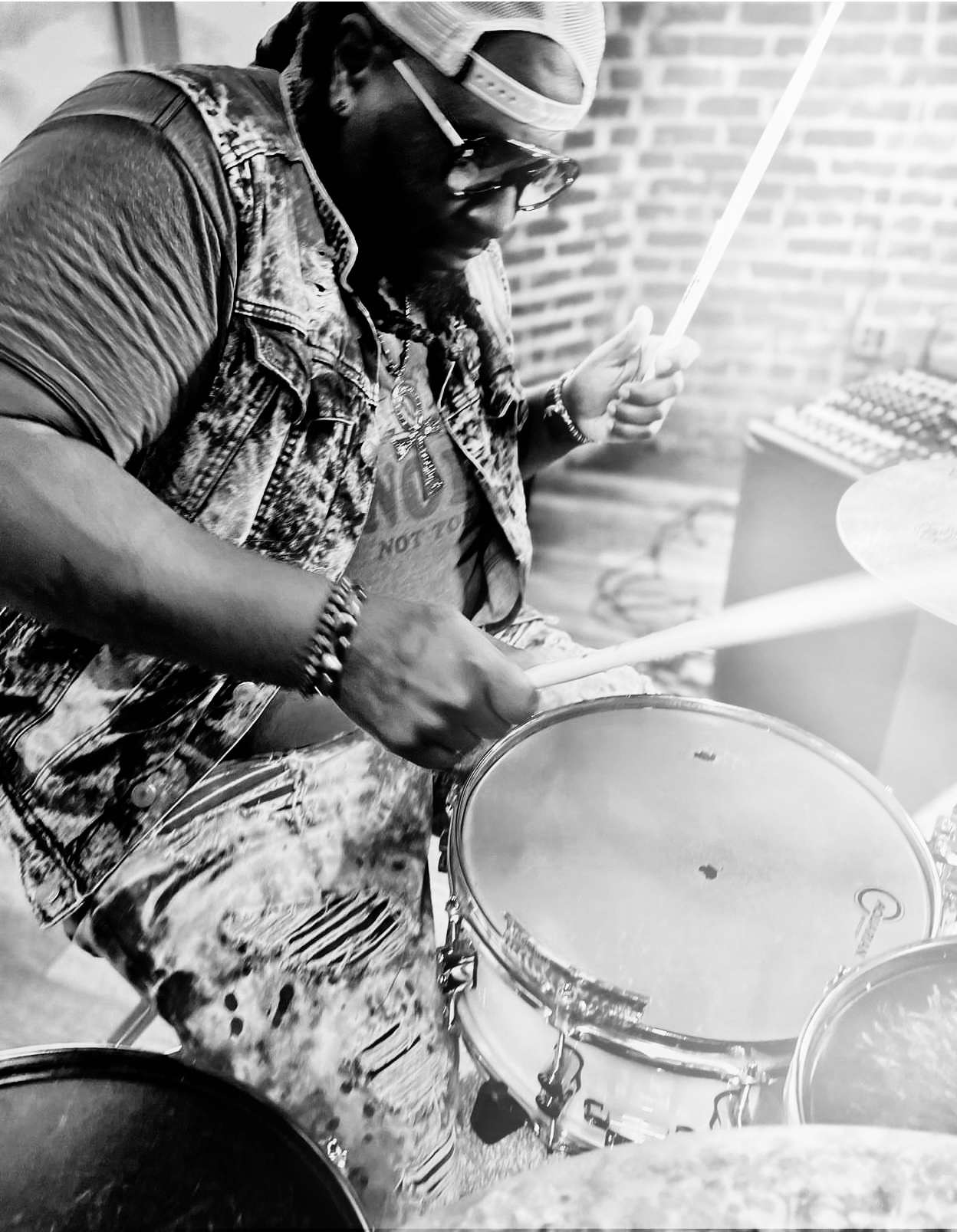 Person playing drum outdoors, wearing glasses, bandana, denim vest, and holding drumsticks.
