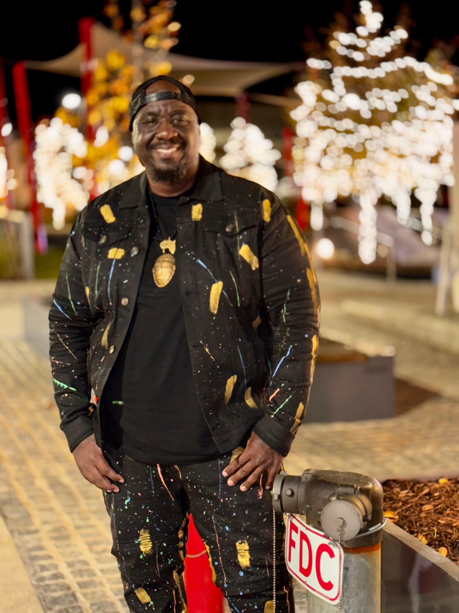 Smiling man wearing black jacket with yellow paint splatters, standing outdoors at night with illuminated trees in background.