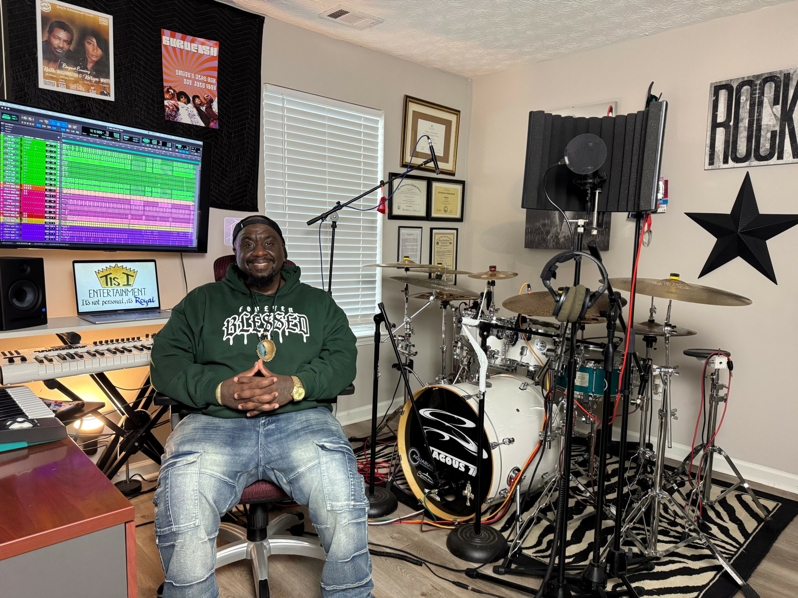 Person sitting in music studio with drums, keyboard, and sound equipment, smiling, wearing green hoodie and jeans.