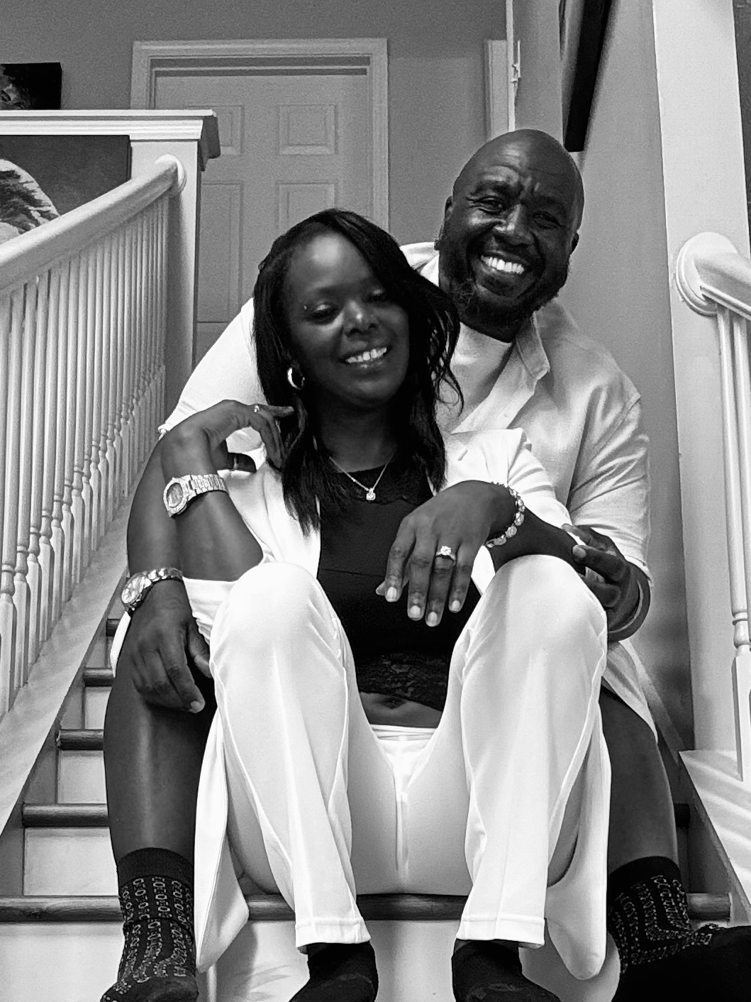 Two people sitting on stairs, smiling, one with arm around the other, in a home interior setting.