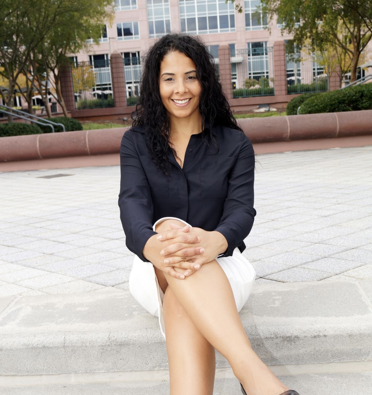 Young woman with curly hair smiling, sitting outdoors on a bench with crossed legs, in front of a building and trees.