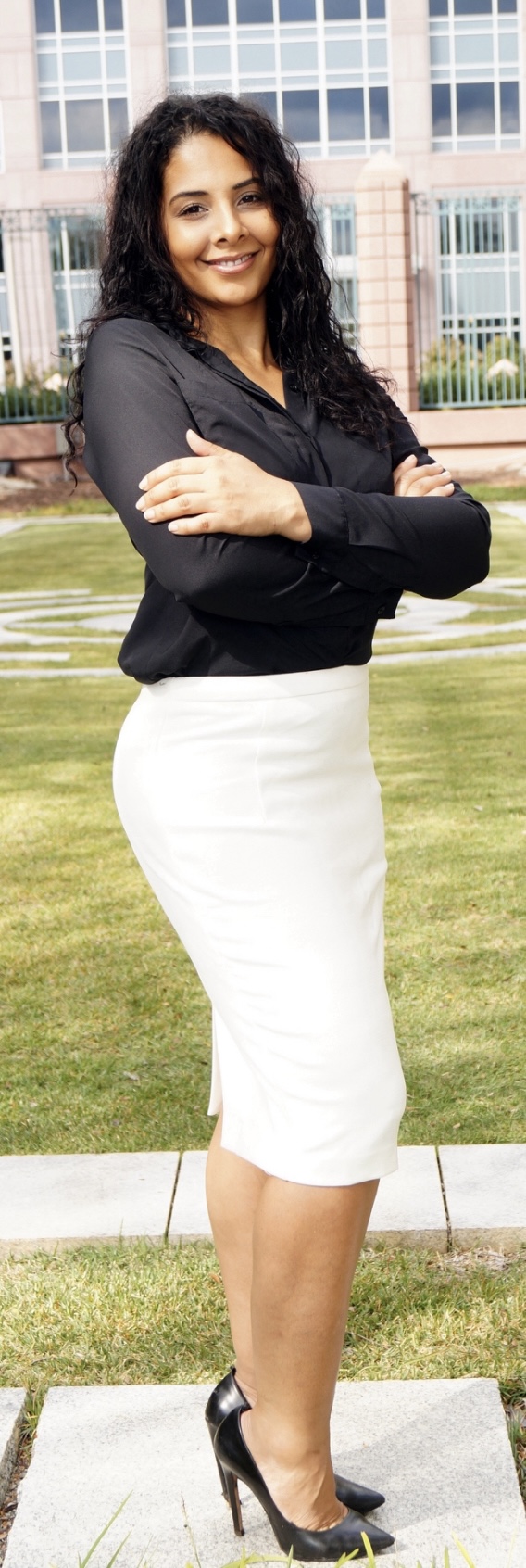 Woman standing outdoors with arms crossed, smiling, in front of a building and grass.