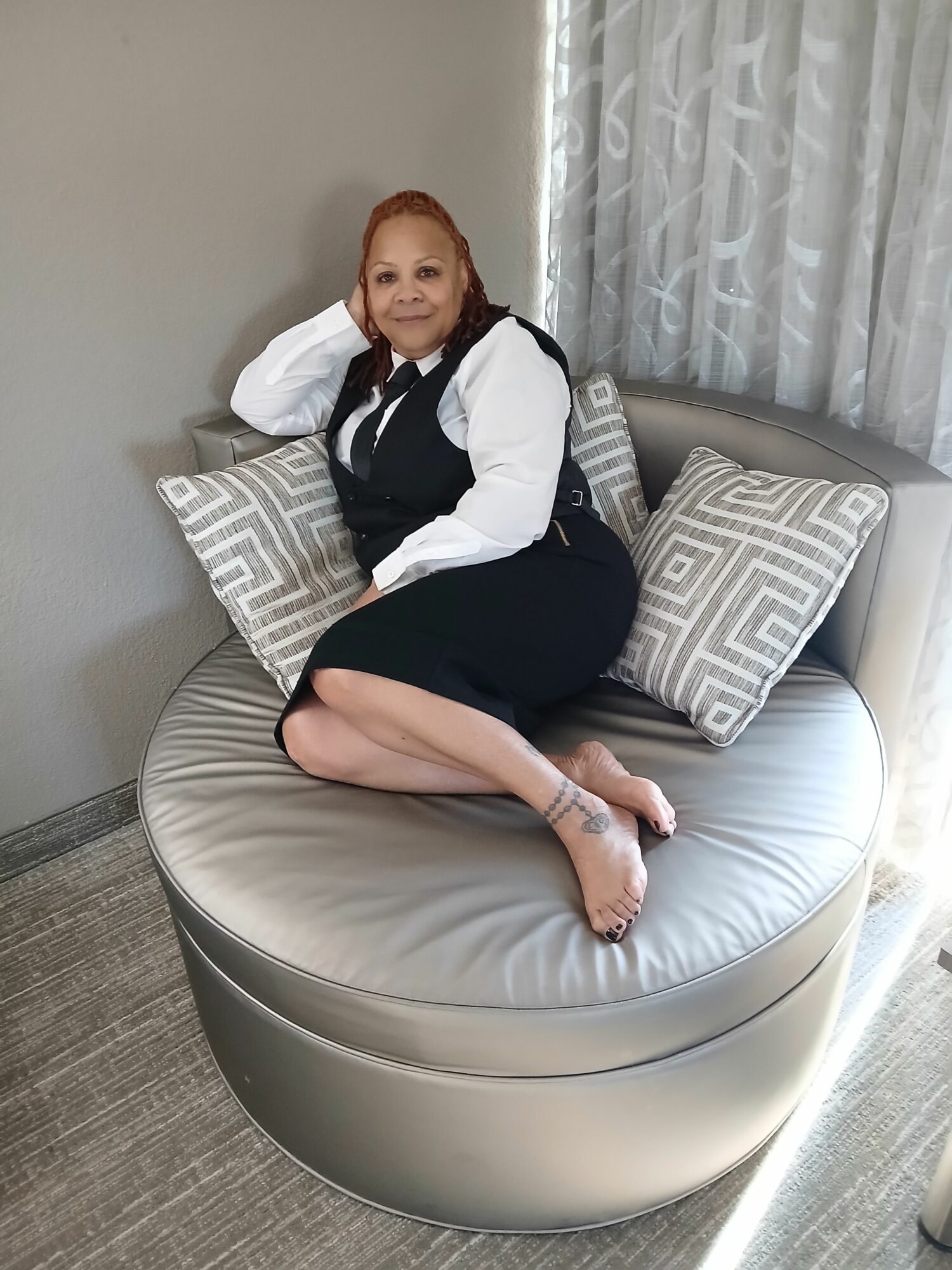 Woman sitting on round bed with pillows, smiling, in a room with curtains and gray walls.