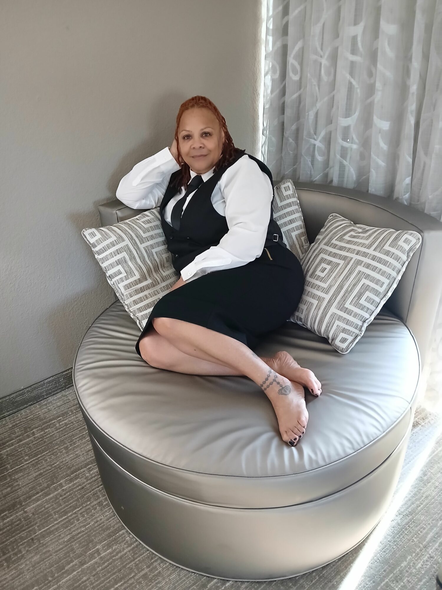 Woman sitting on a round sofa with pillows, resting her head on her hand, smiling, in a room with curtains.