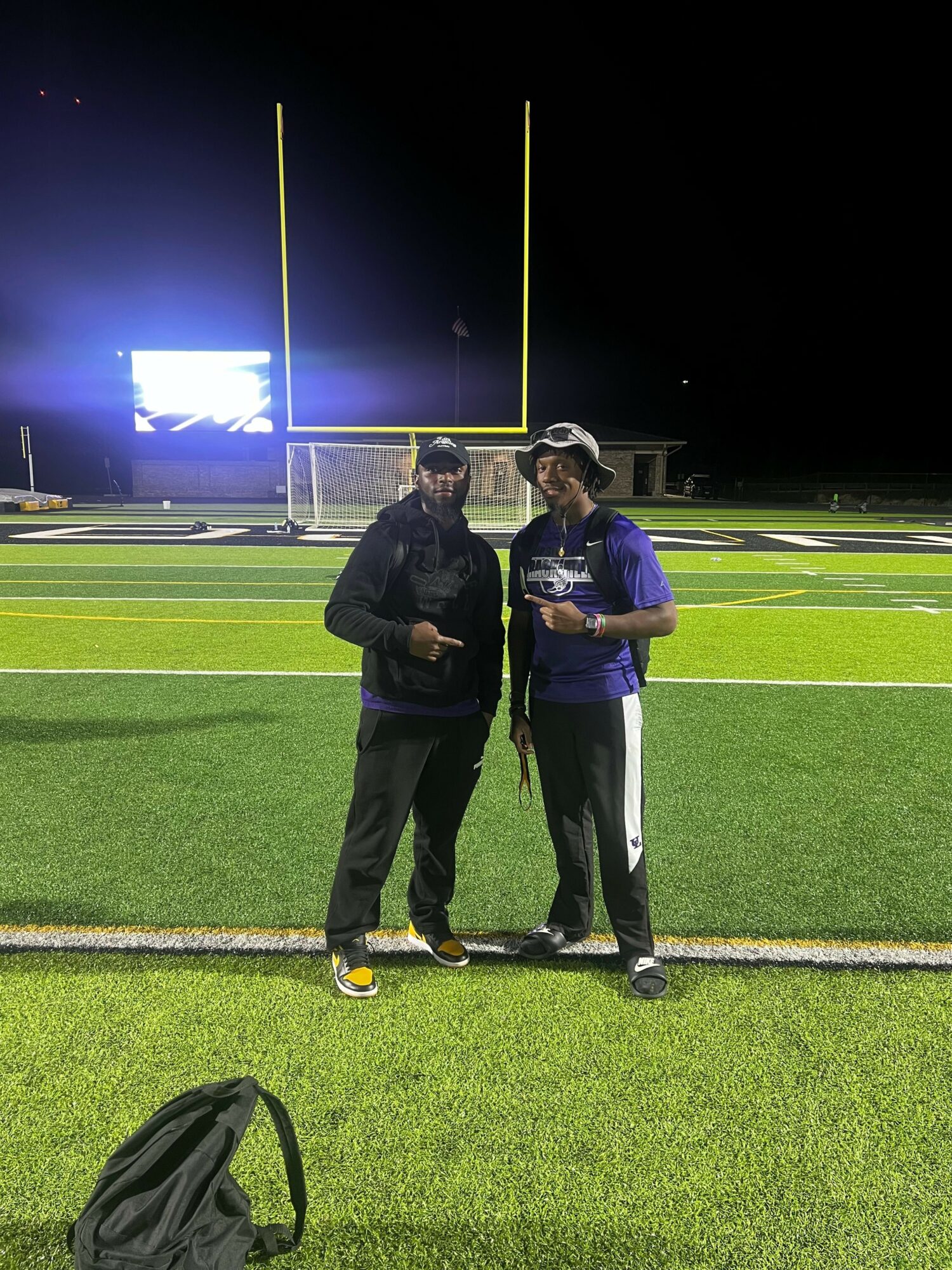 Two people standing on a football field at night, holding drinks, with goalposts and a scoreboard in the background.