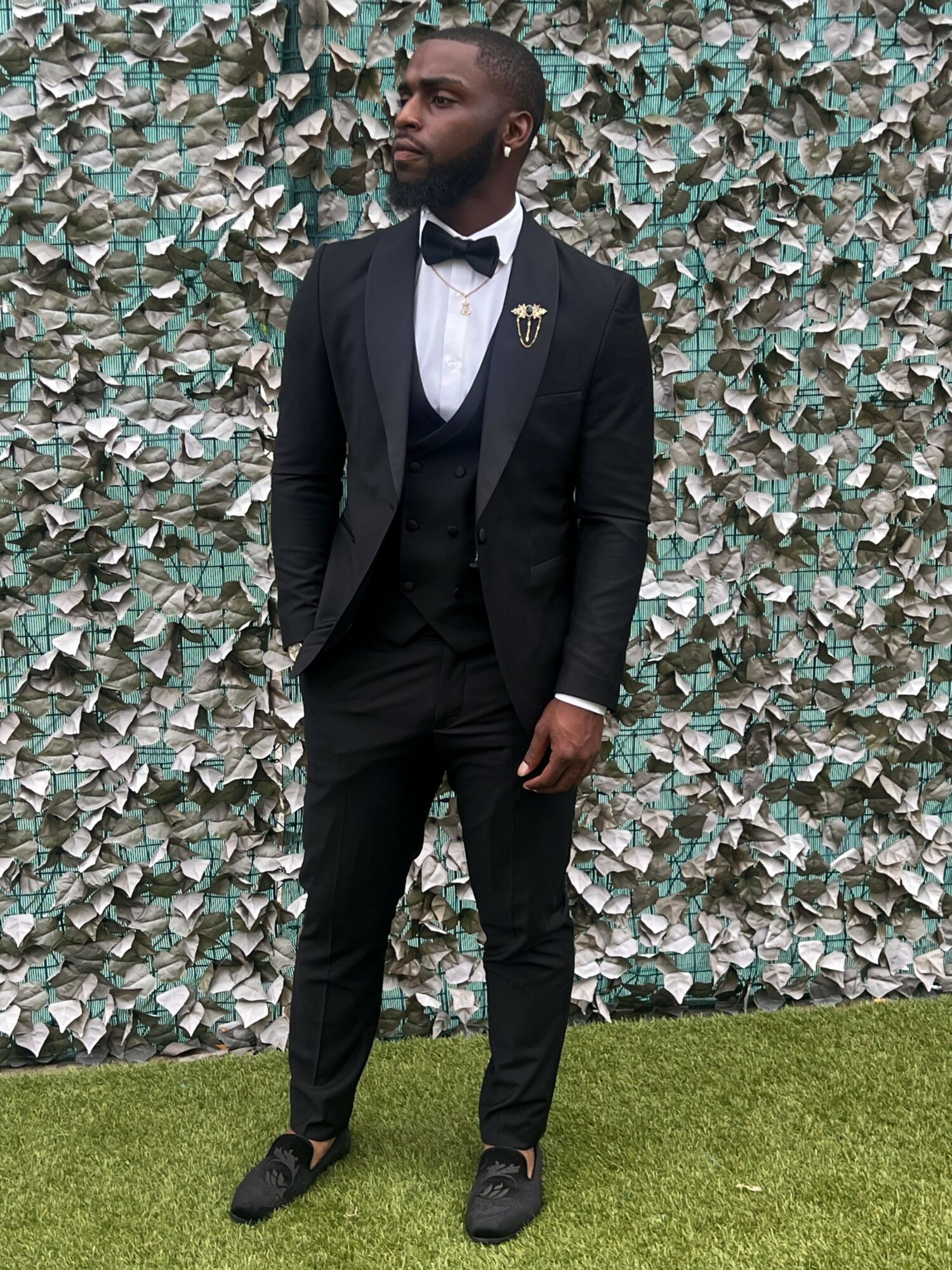 Man in black tuxedo with bow tie standing outdoors against a leafy background.