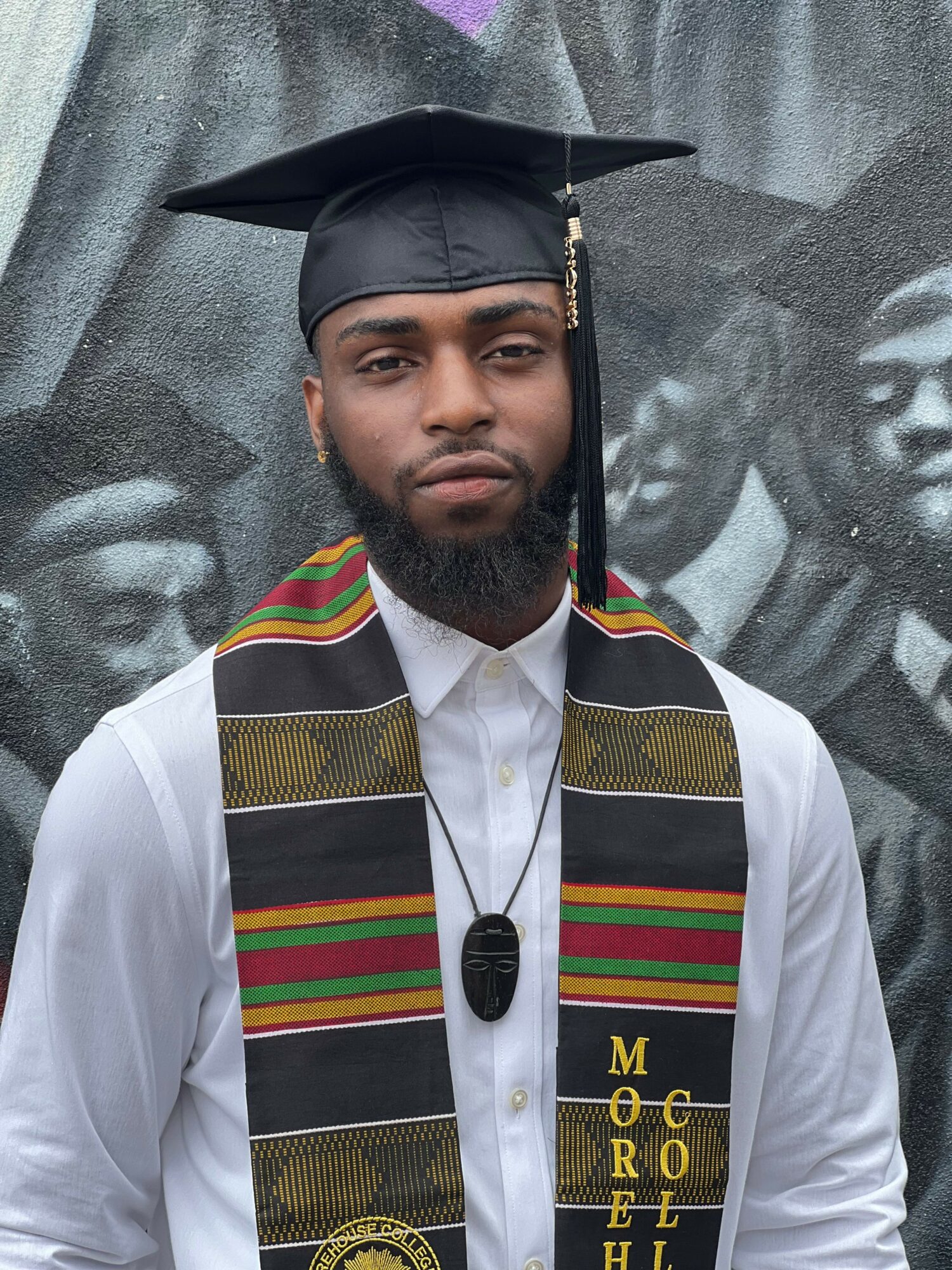 Young man in graduation cap and gown with colorful sash, standing in front of a black-and-white photo backdrop.