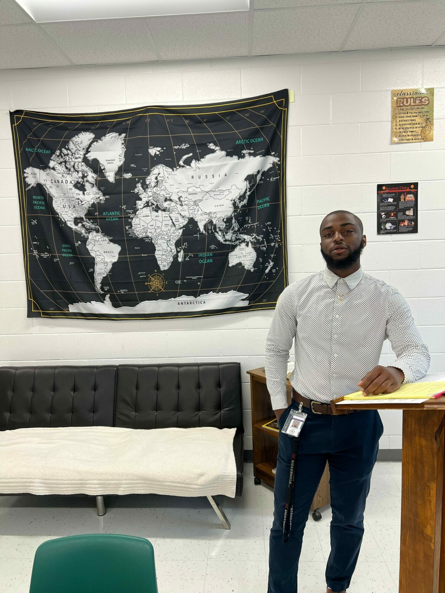 Man standing next to a world map on a wall, in a room with white walls and furniture.