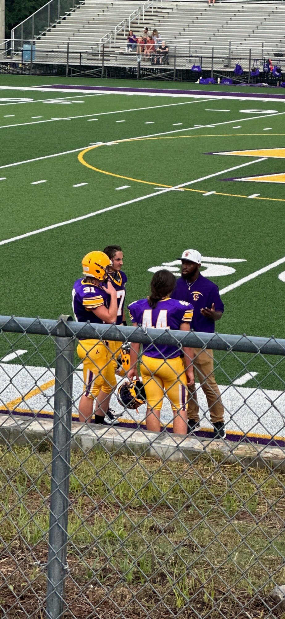 Three football players and a coach standing on the sideline of a football field, with empty bleachers in the background.
