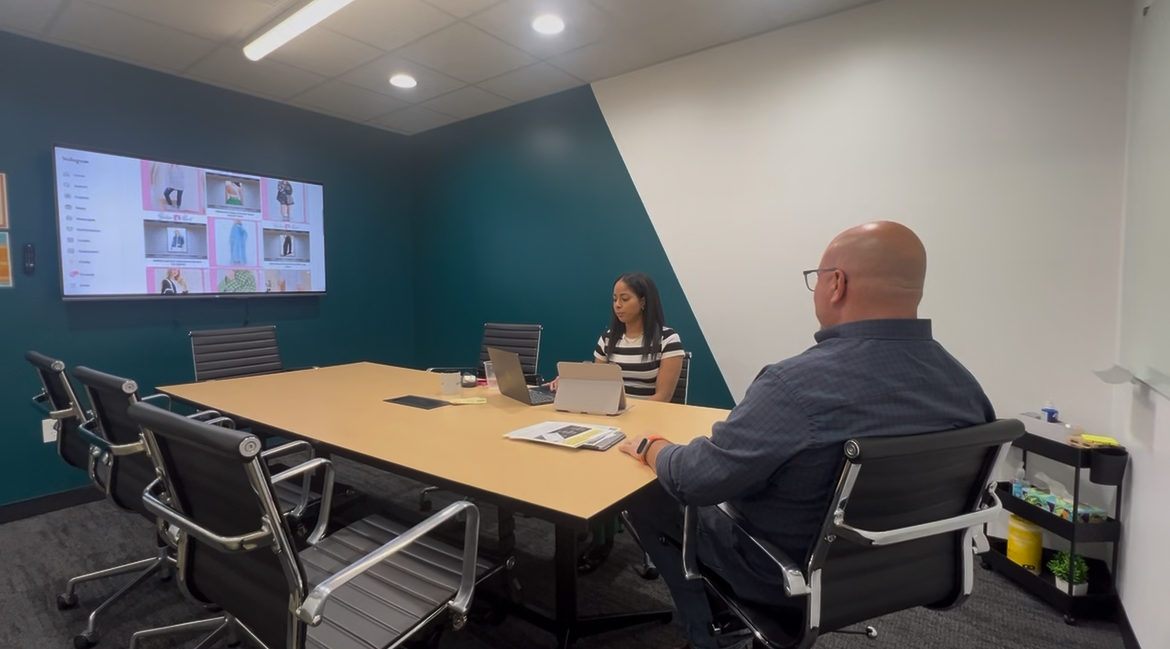 Two people sit at a conference table in a modern meeting room with a large screen displaying multiple images.