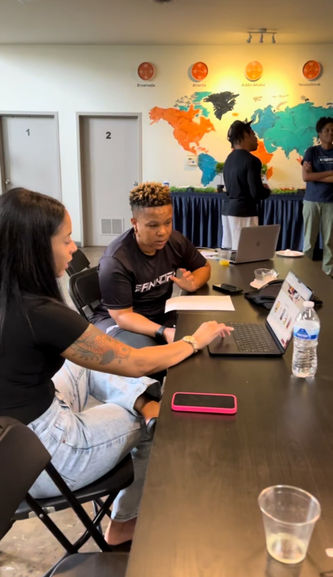 Two people seated at a table with laptops, water bottle, and phone, in a room with a world map on the wall.