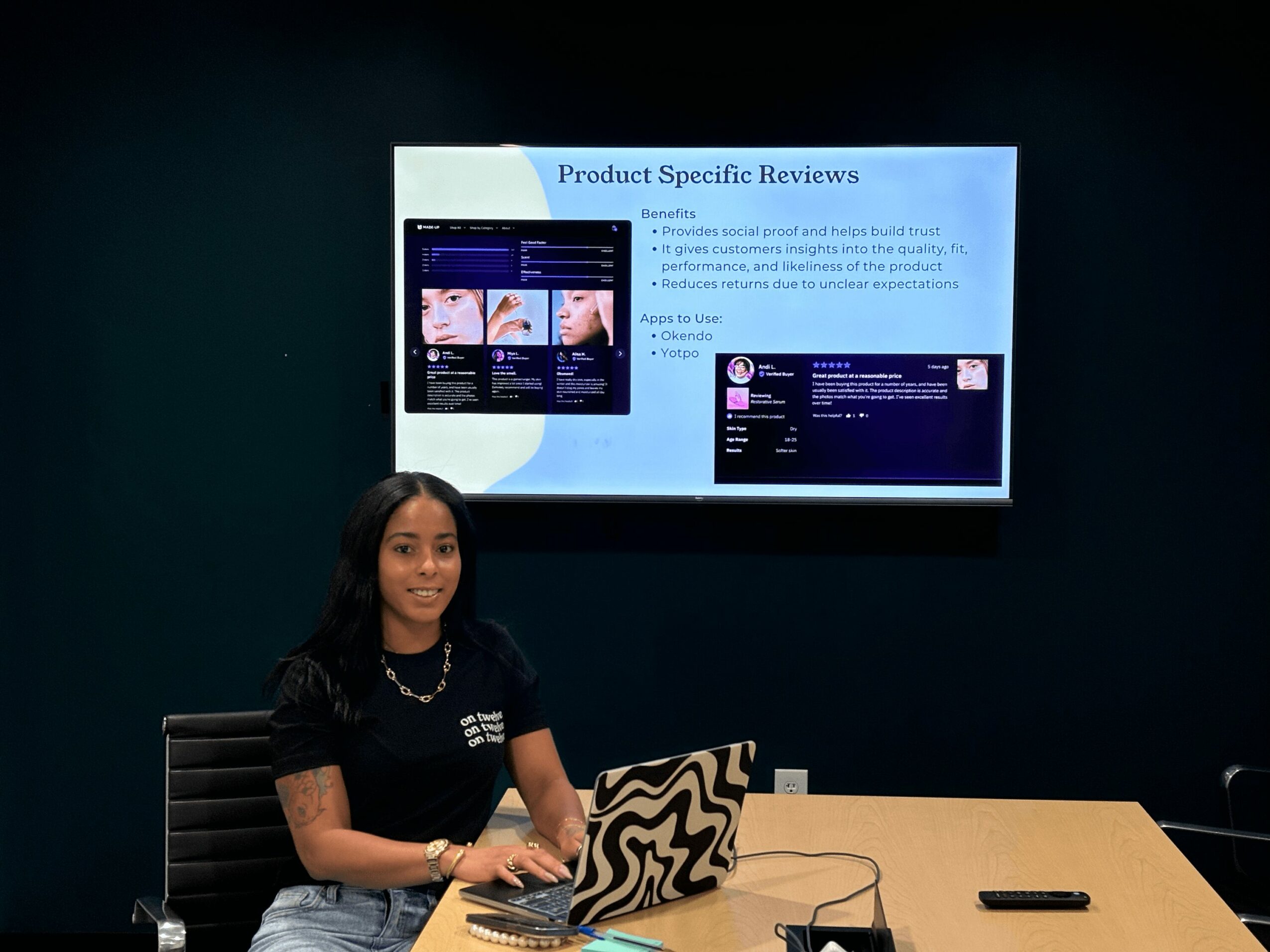 Woman sitting at a conference table with a laptop, presenting a slide on a screen behind her.