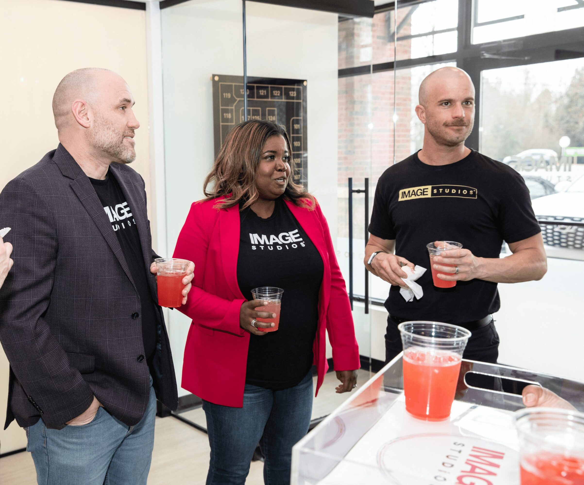 Three people standing indoors, holding drinks, near a table with a large container of red beverage, large windows in background.