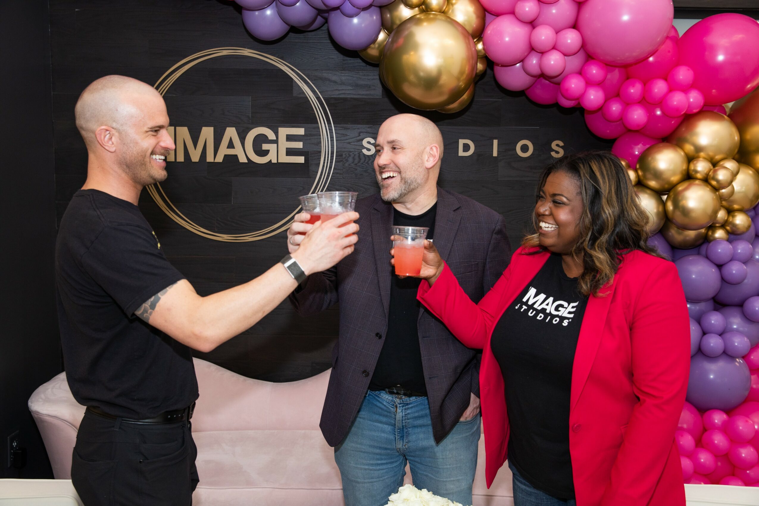 Three people smiling and toasting with drinks at a celebration event with colorful balloon decorations in the background.