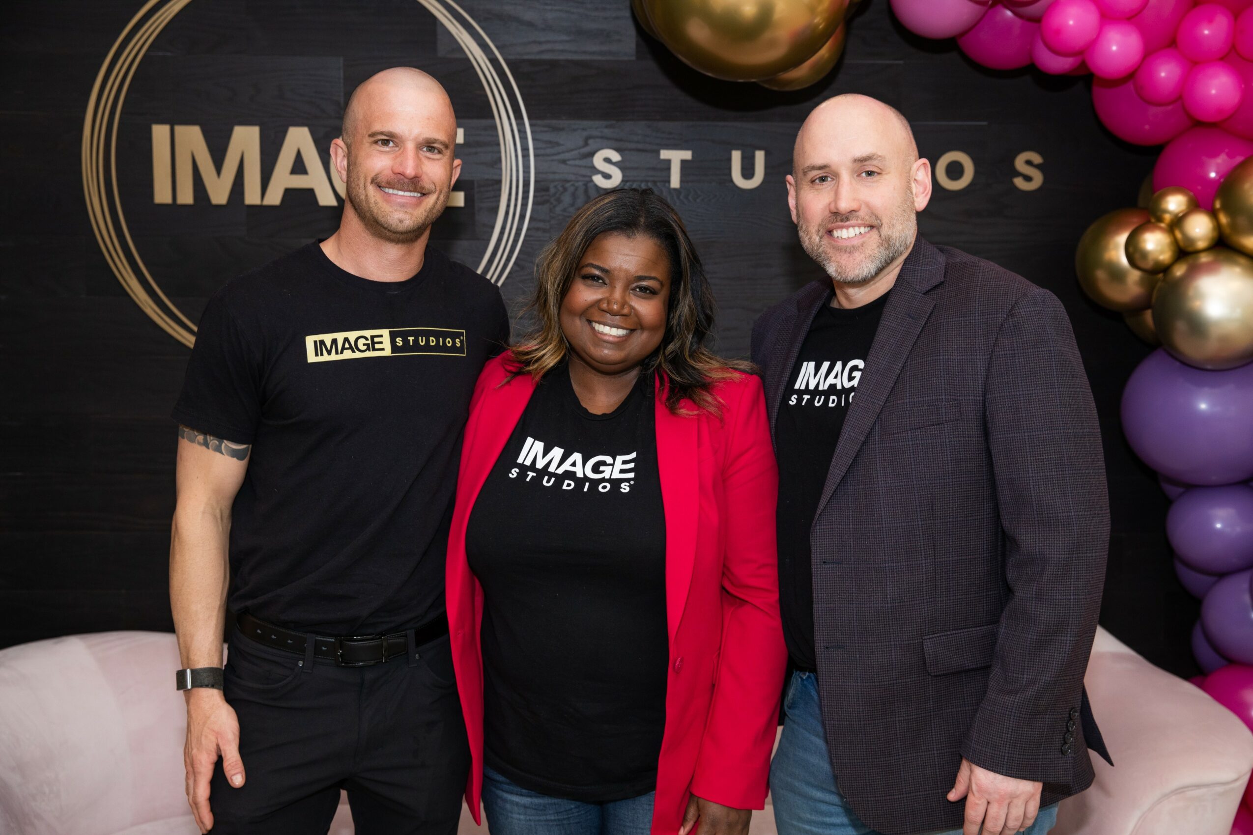 Three people standing together, smiling, in front of a decorated backdrop with balloons and a logo, indoors.