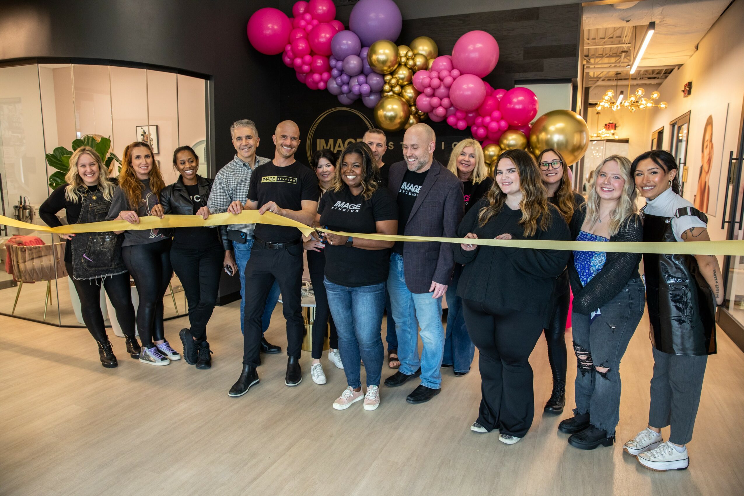 Group of people standing indoors with colorful balloons overhead, holding a yellow ribbon for a celebration or event.