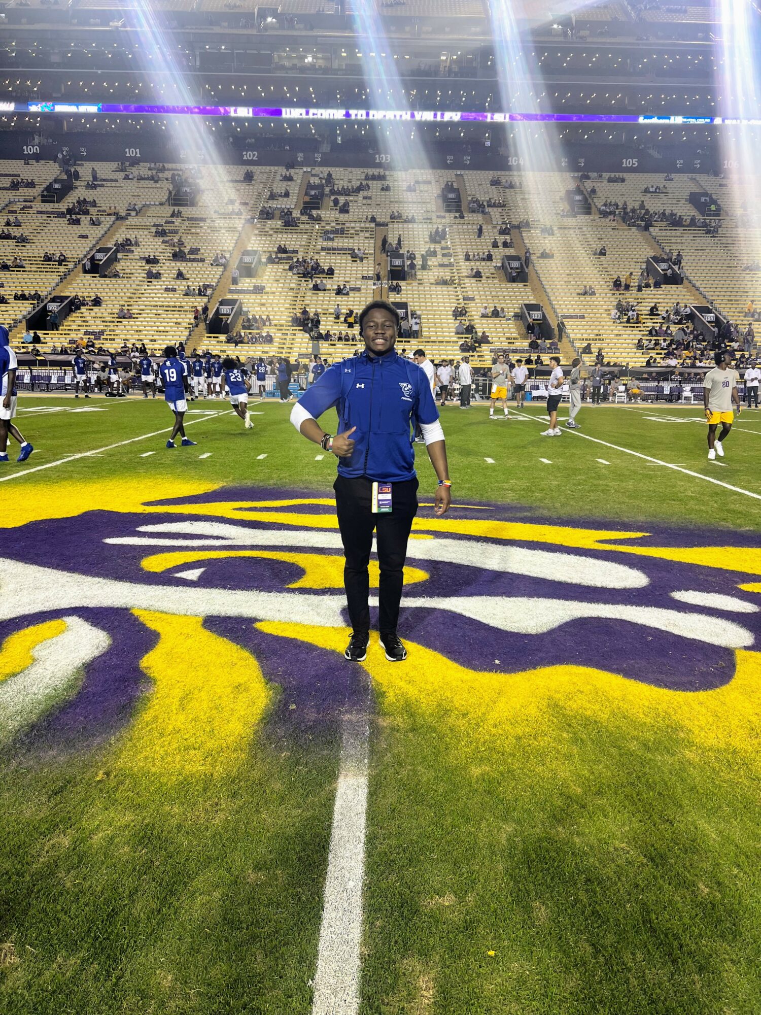 Person standing on a football field with stadium seating and players in the background.