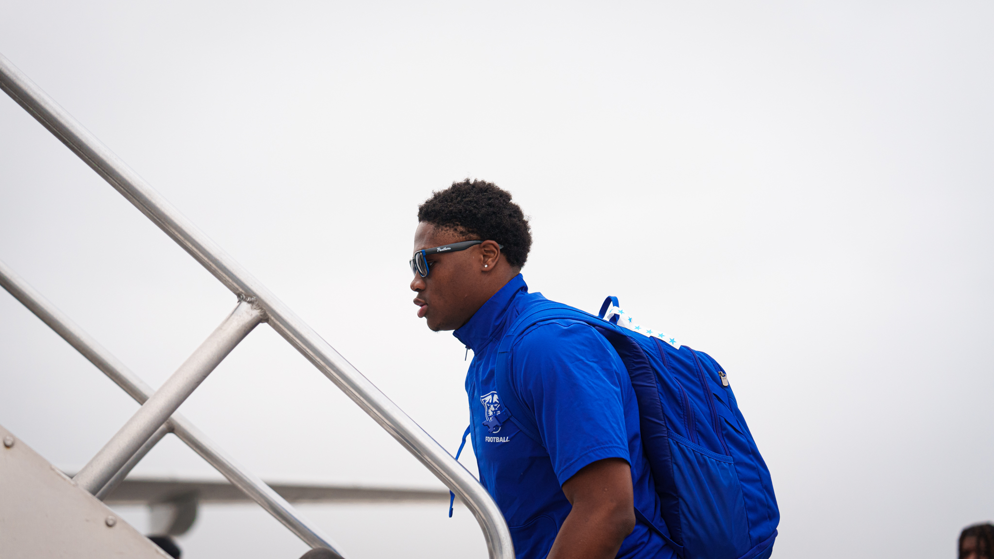 Young man with sunglasses and a blue backpack walking up stairs against a plain white background.