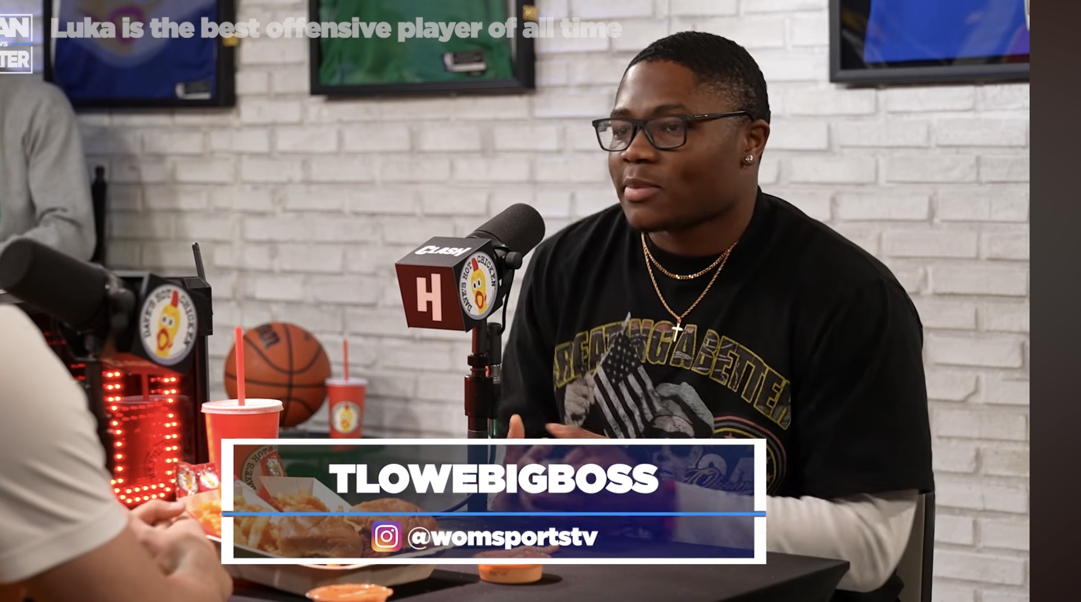 Young man with glasses and black T-shirt sitting at a table during an interview, with a white brick wall background.