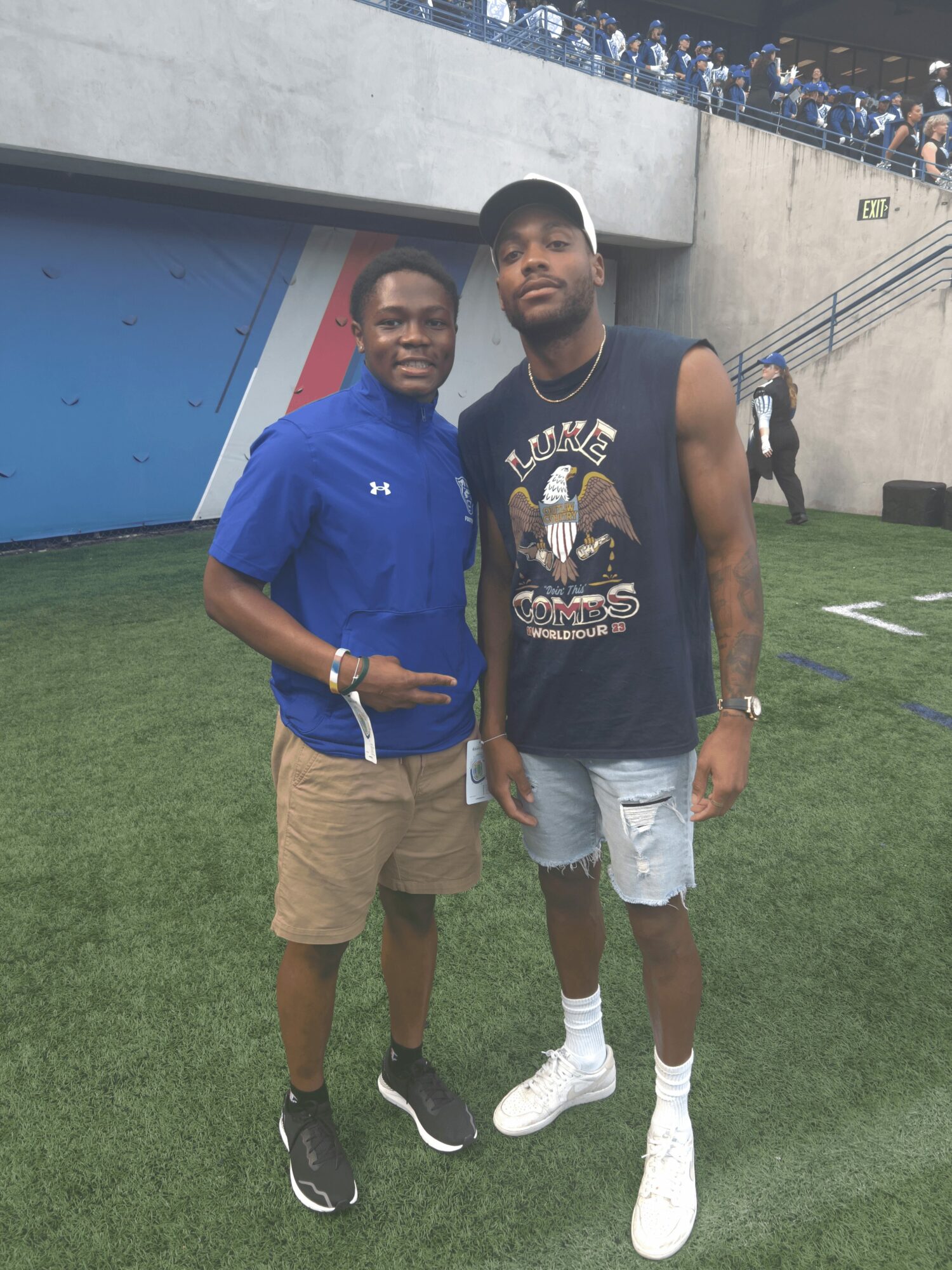 Two young men standing on a grassy field, one in a blue jacket and beige shorts, the other in a black sleeveless shirt and ripped shorts.