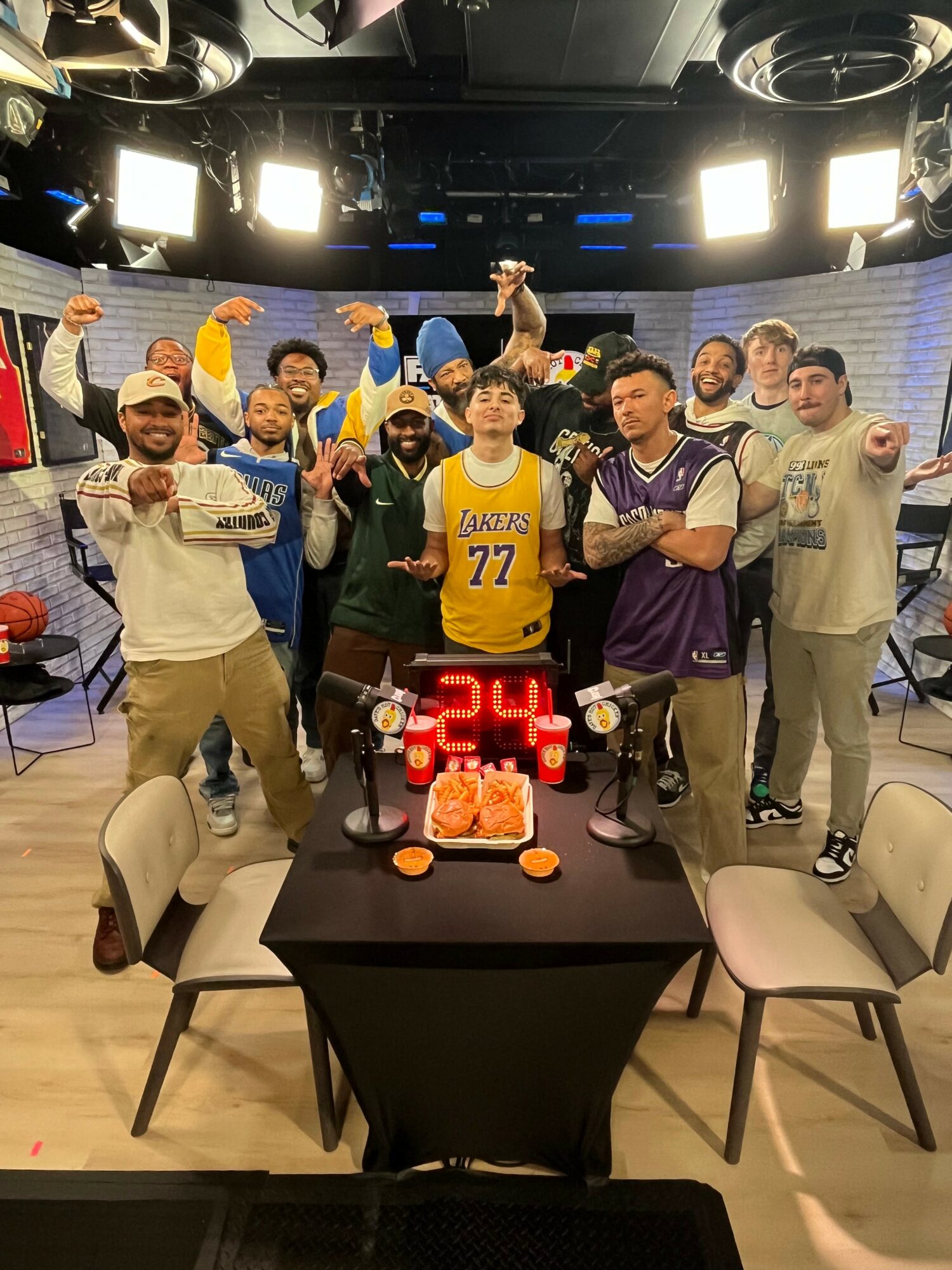 Group of people celebrating around a table with snacks and a digital timer, some raising fists, in a studio setting.