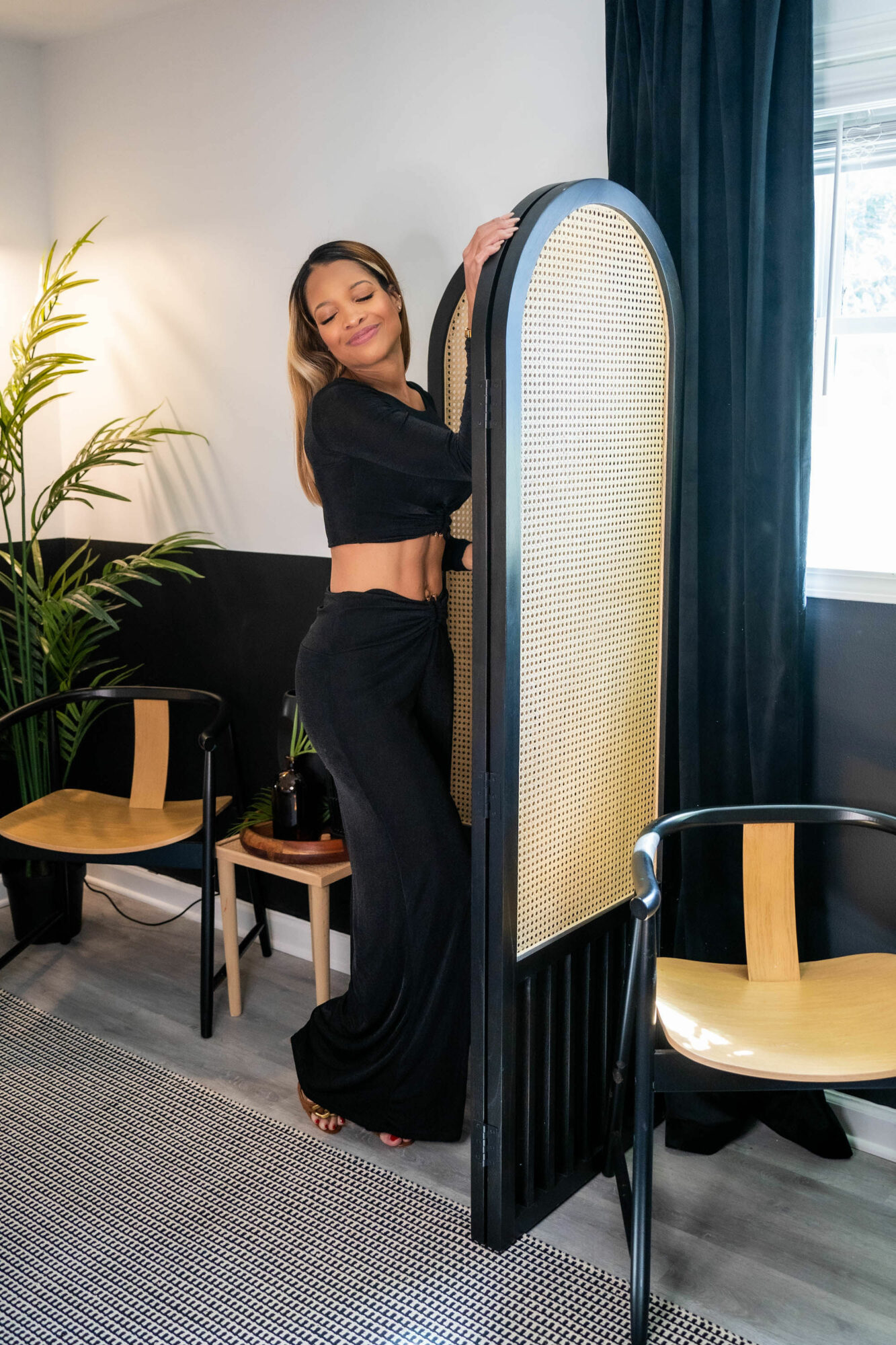 Woman standing behind a folding room divider, smiling, in a room with chairs, a plant, and a window with curtains.