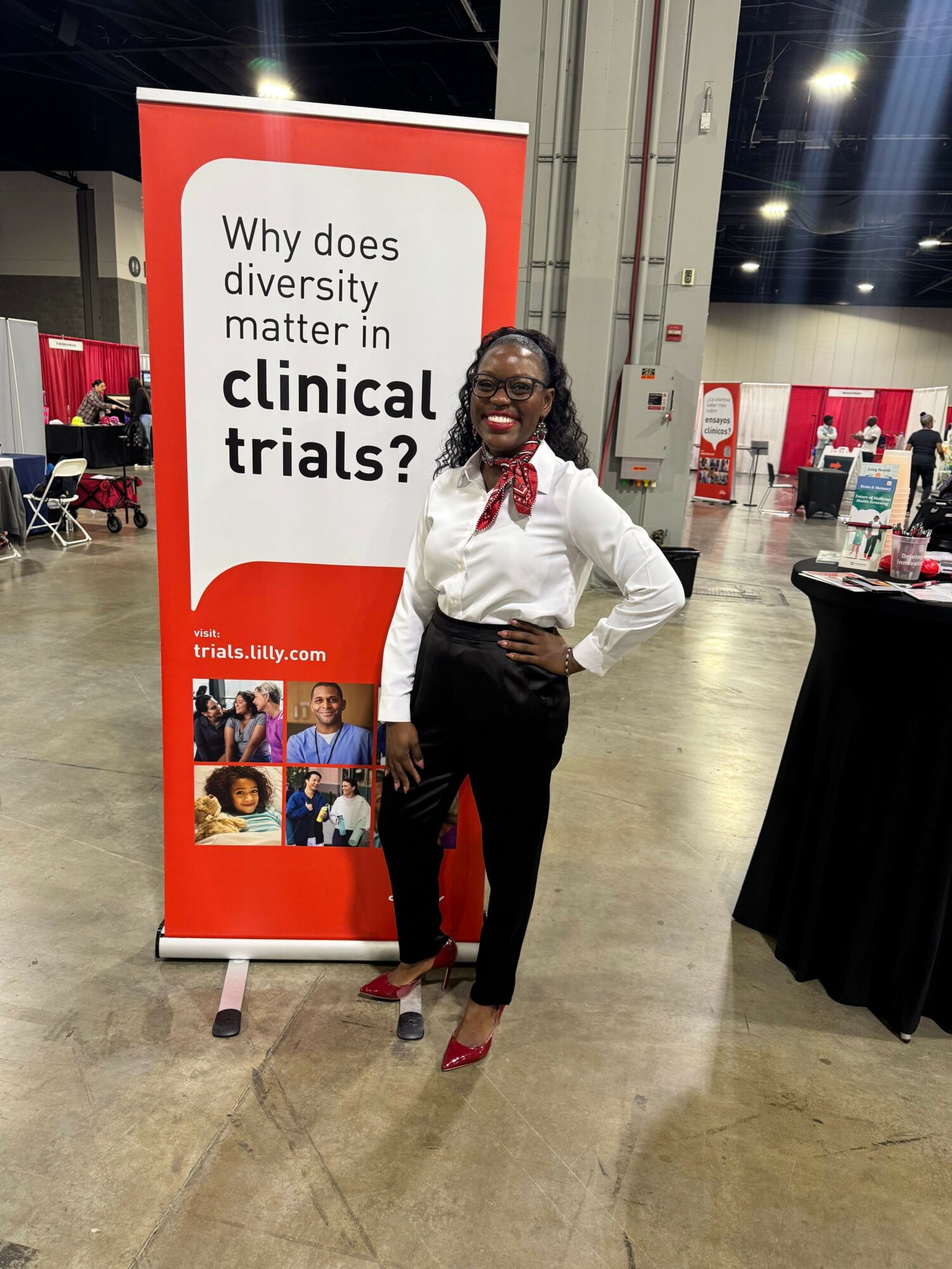 Woman standing next to a red and white banner at an indoor event, smiling with hands on hips.