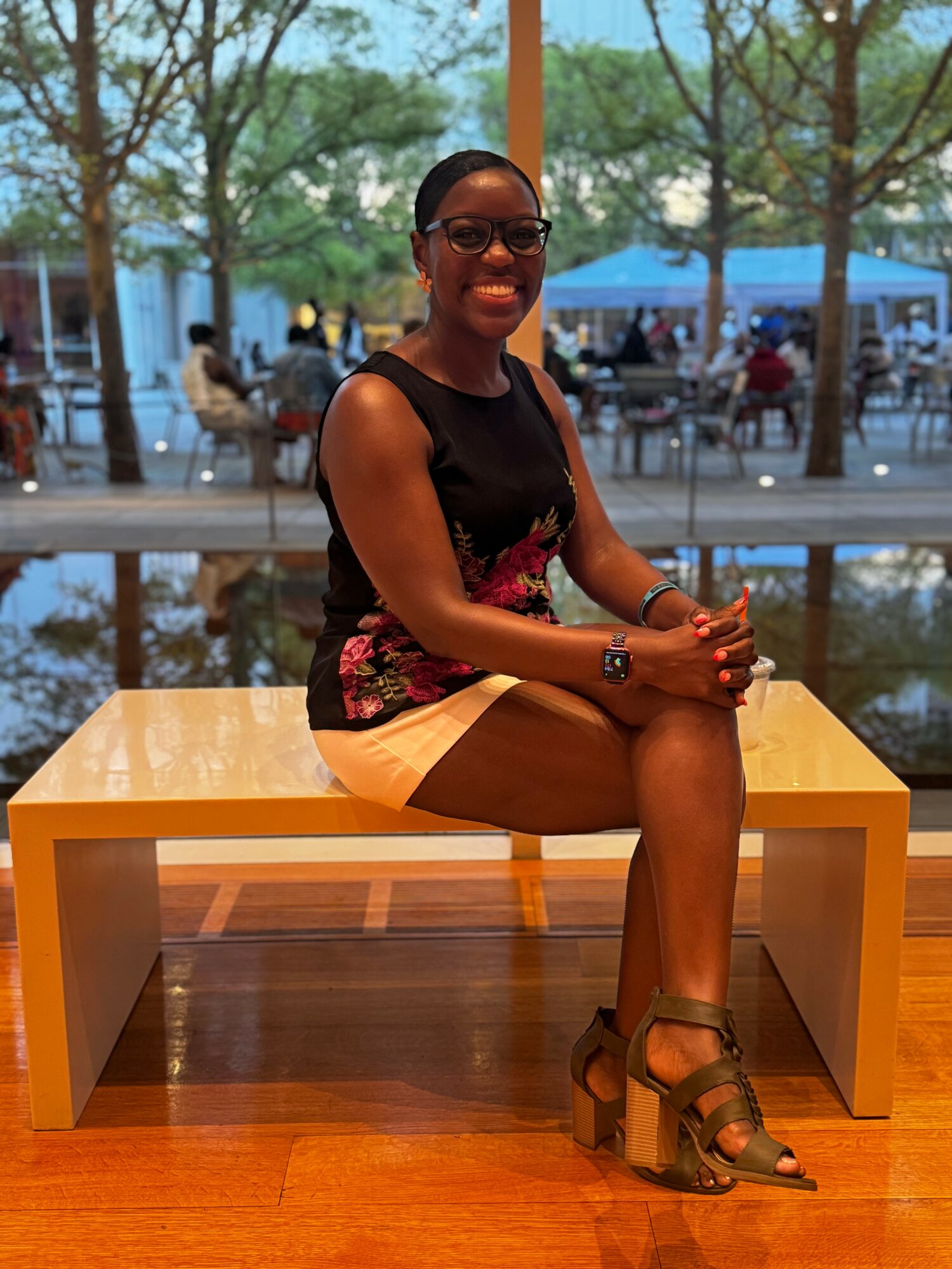 Woman sitting on a yellow bench indoors, smiling, wearing glasses, a black sleeveless top, and beige shorts.