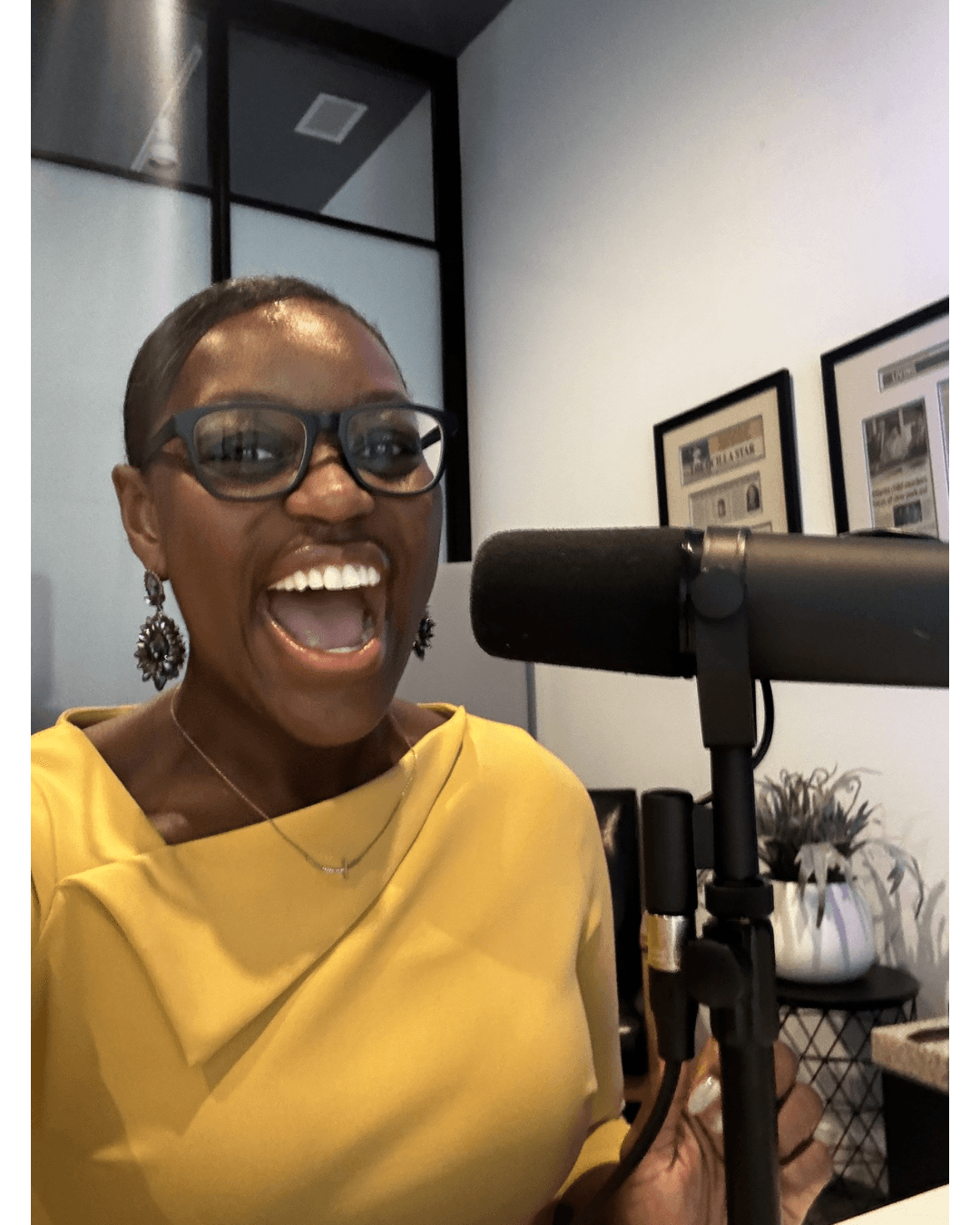 Woman with glasses smiling and speaking into a microphone in a room with framed pictures and plants.