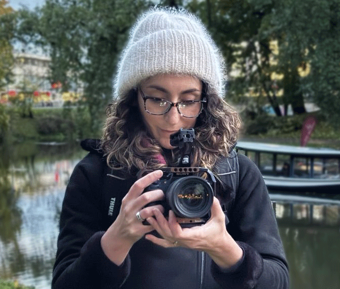 Person wearing glasses and a white knit hat takes a photo outdoors near water with trees and a boat.
