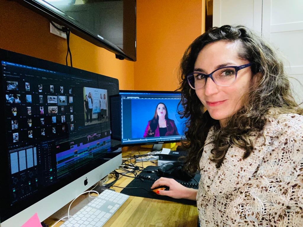 Woman with glasses and curly hair working at desk with two monitors, one showing video editing software, the other a video call.