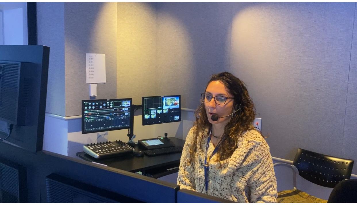Woman with glasses and curly hair working at a computer station with multiple monitors, in a control room.