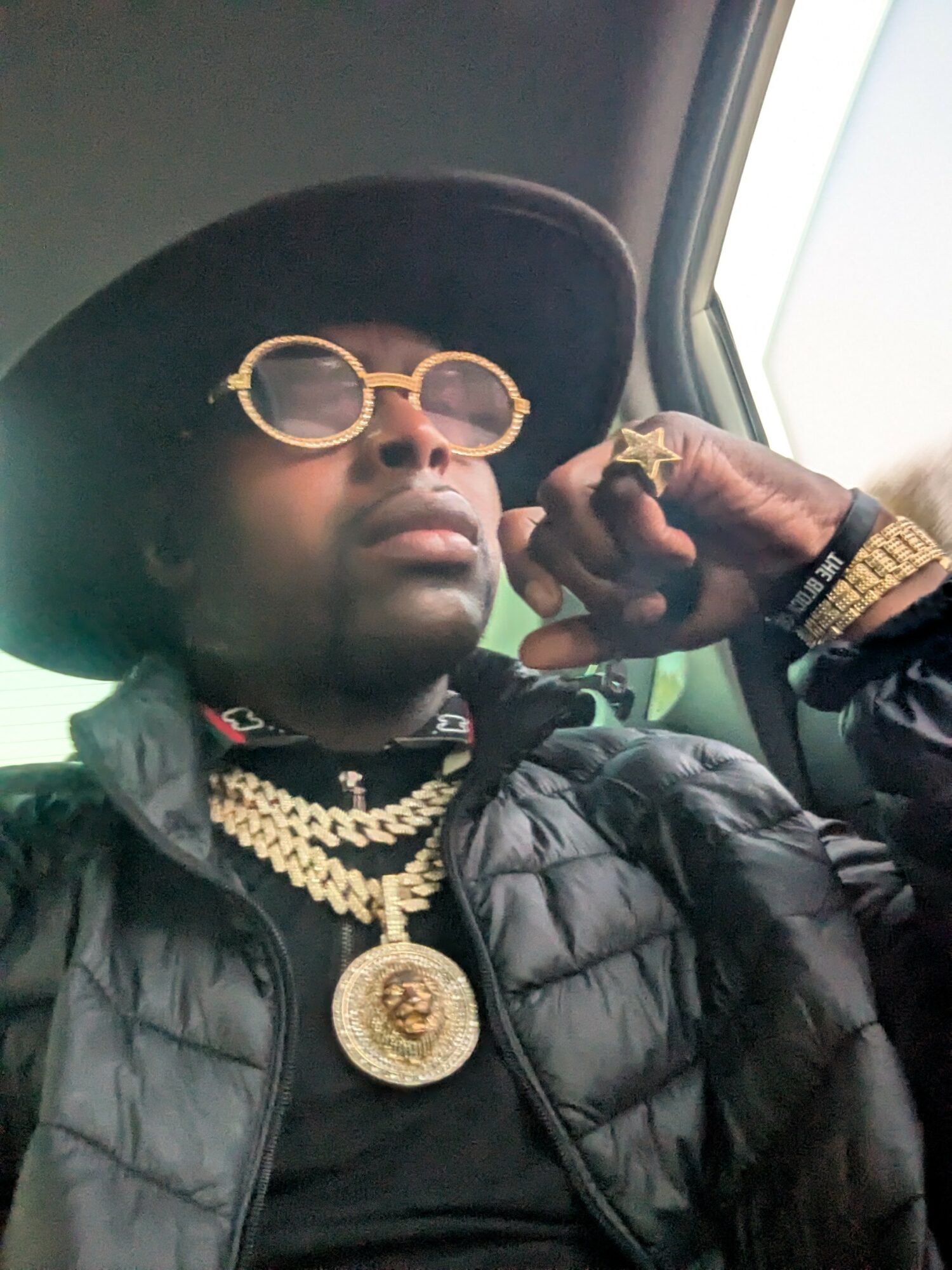 Person wearing large hat, sunglasses, multiple gold chains, rings, and bracelets, sitting inside a vehicle.