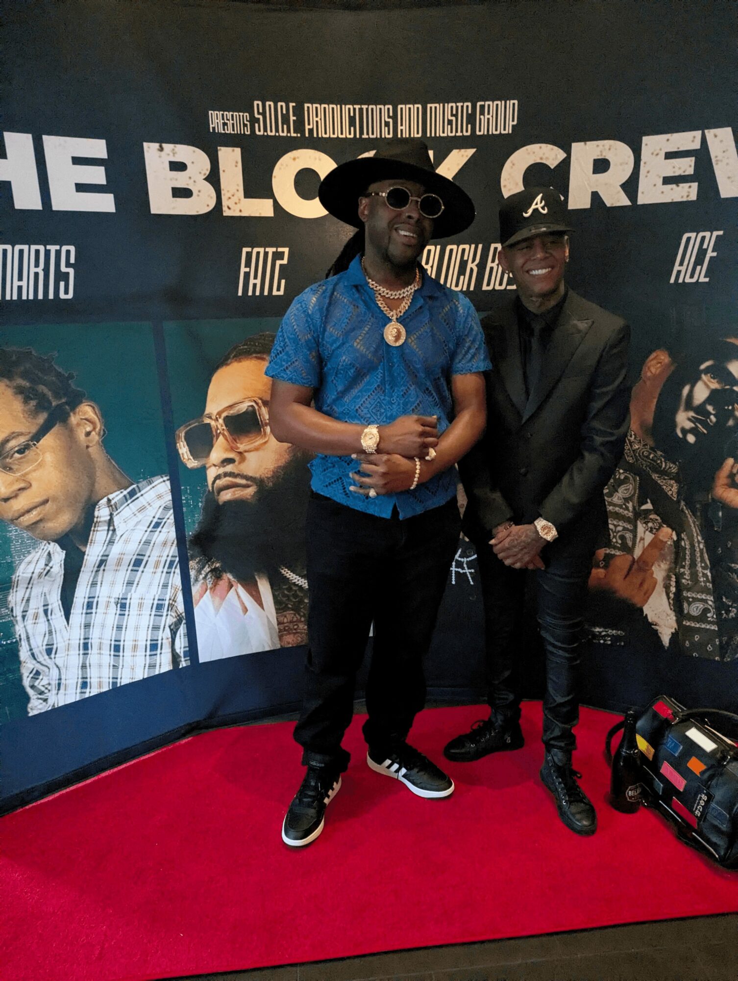 Two men stand on a red carpet in front of a promotional backdrop, smiling and posing for the photo.