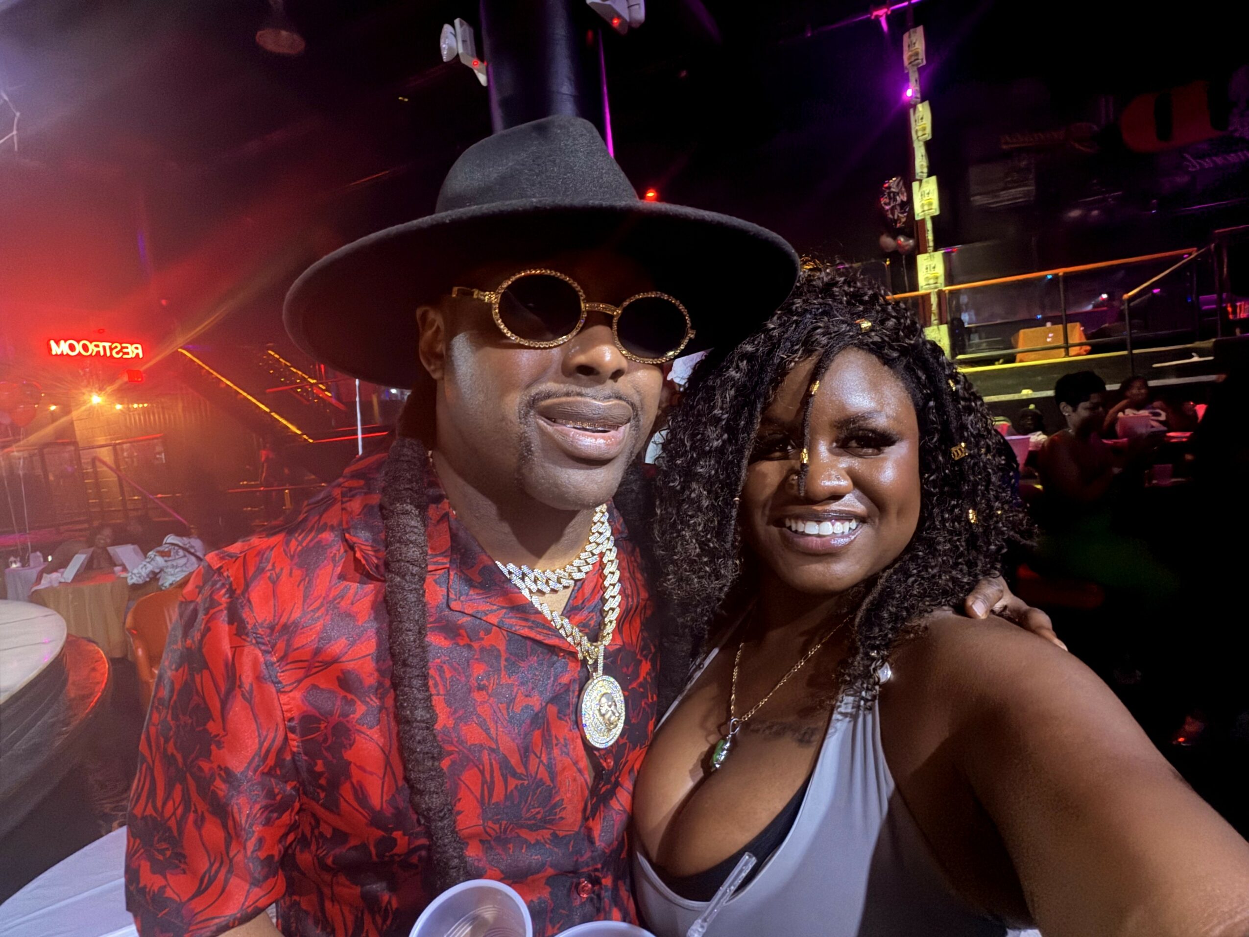 Two people smiling at a party, one wearing a hat, sunglasses, and red patterned shirt, the other in a gray top.