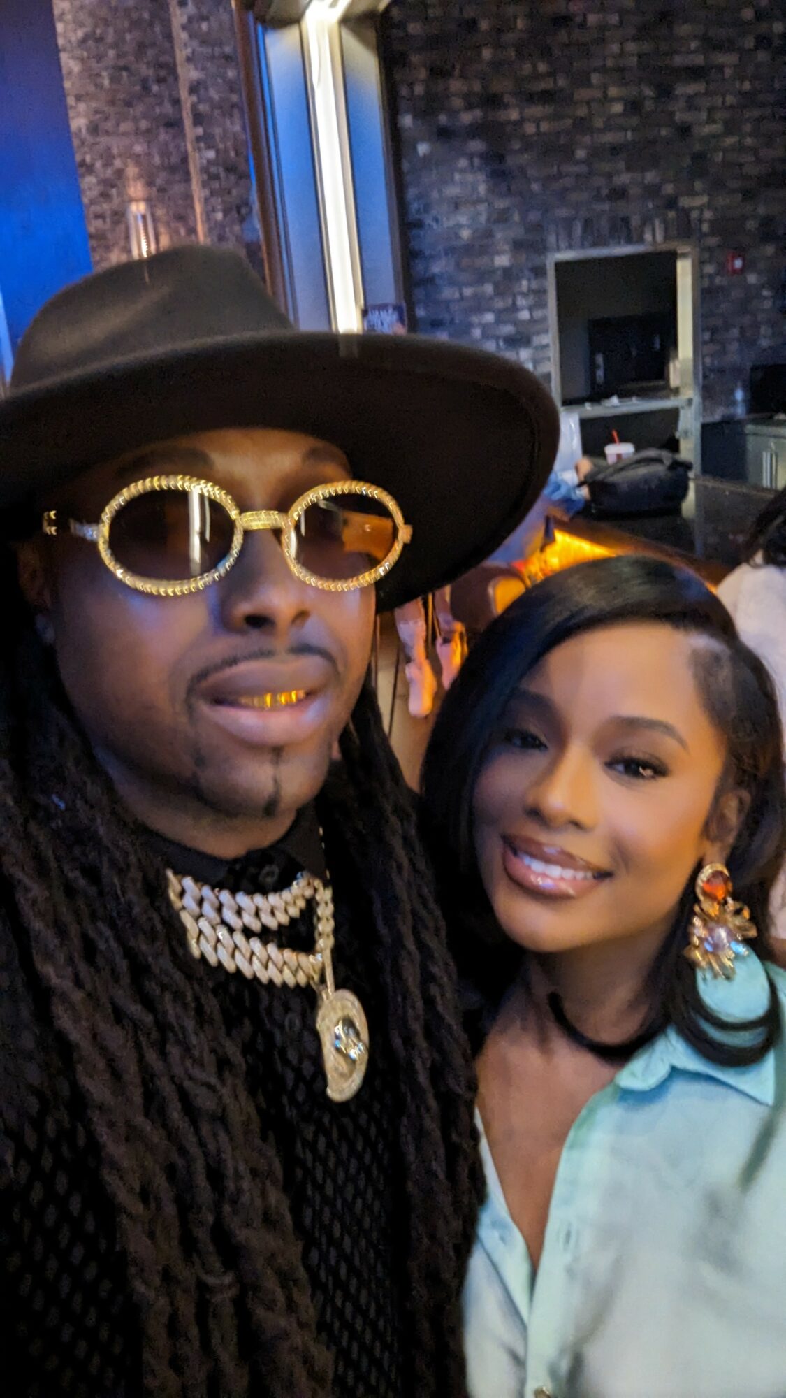 Two people smiling, one wearing large sunglasses, hat, and jewelry, the other with earrings and a light blue top.