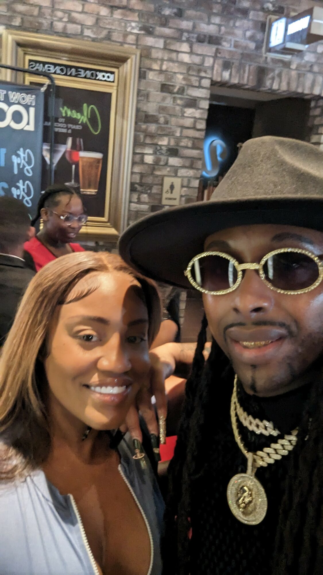 Man wearing hat and sunglasses with gold jewelry, woman smiling, people in background, brick wall, window, and sign behind them.
