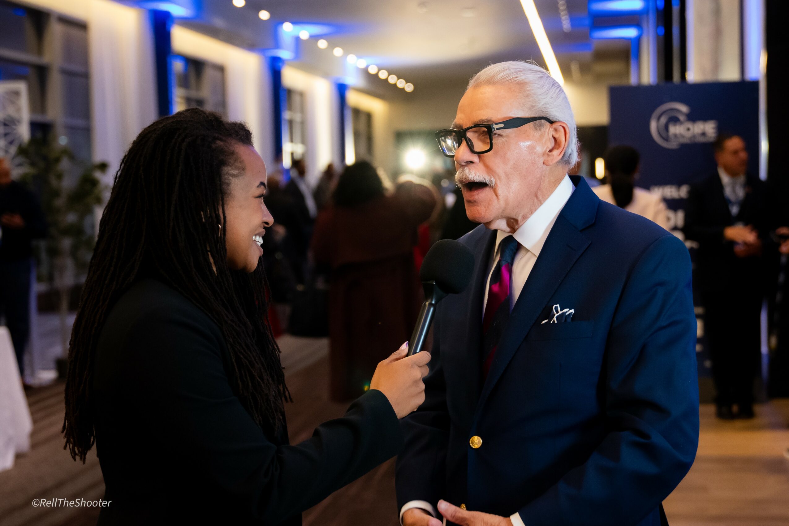 Young woman interviewing an older man with glasses and a suit at an indoor event.