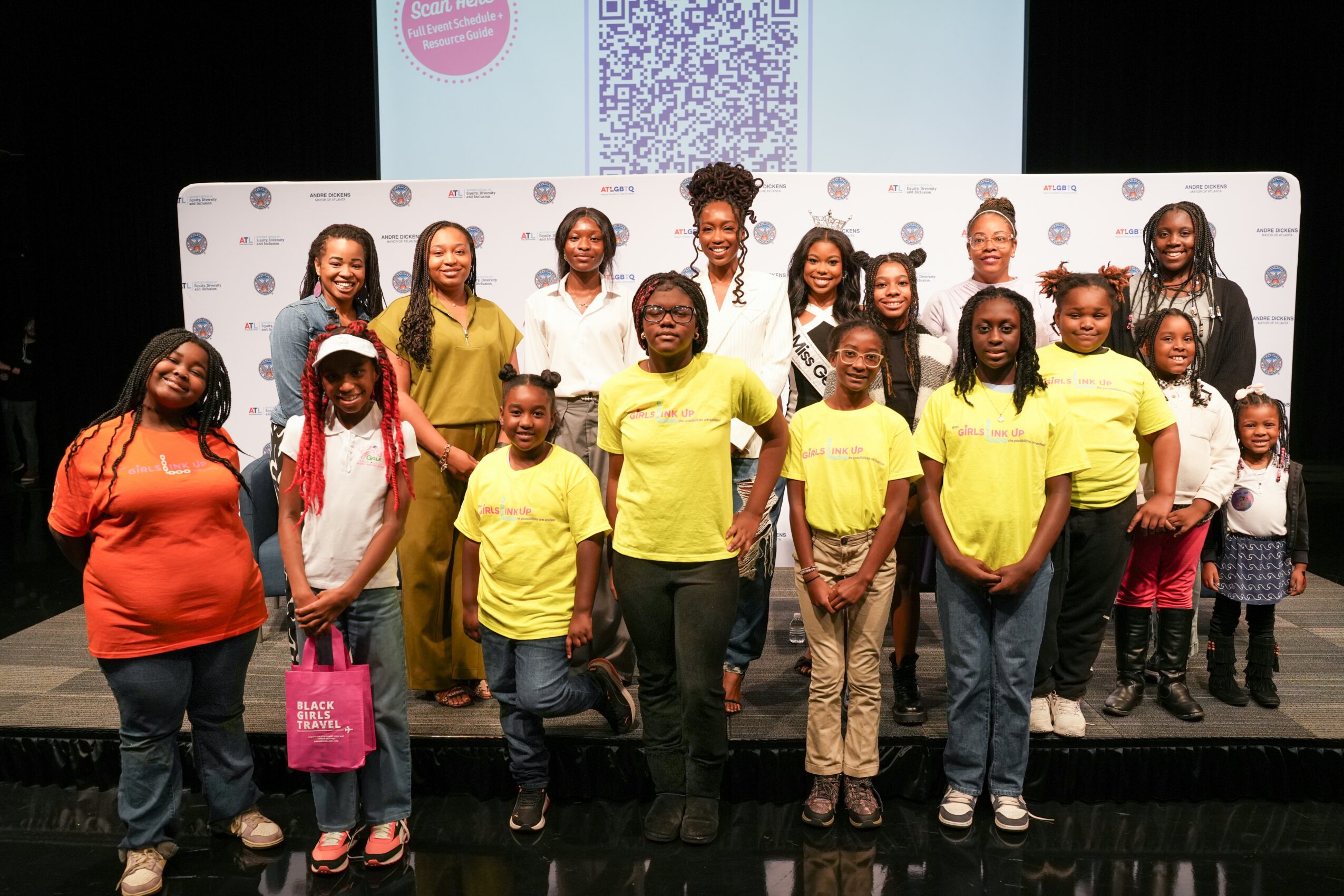 Group of children and adults standing on stage in front of a backdrop and screen, smiling for a photo.