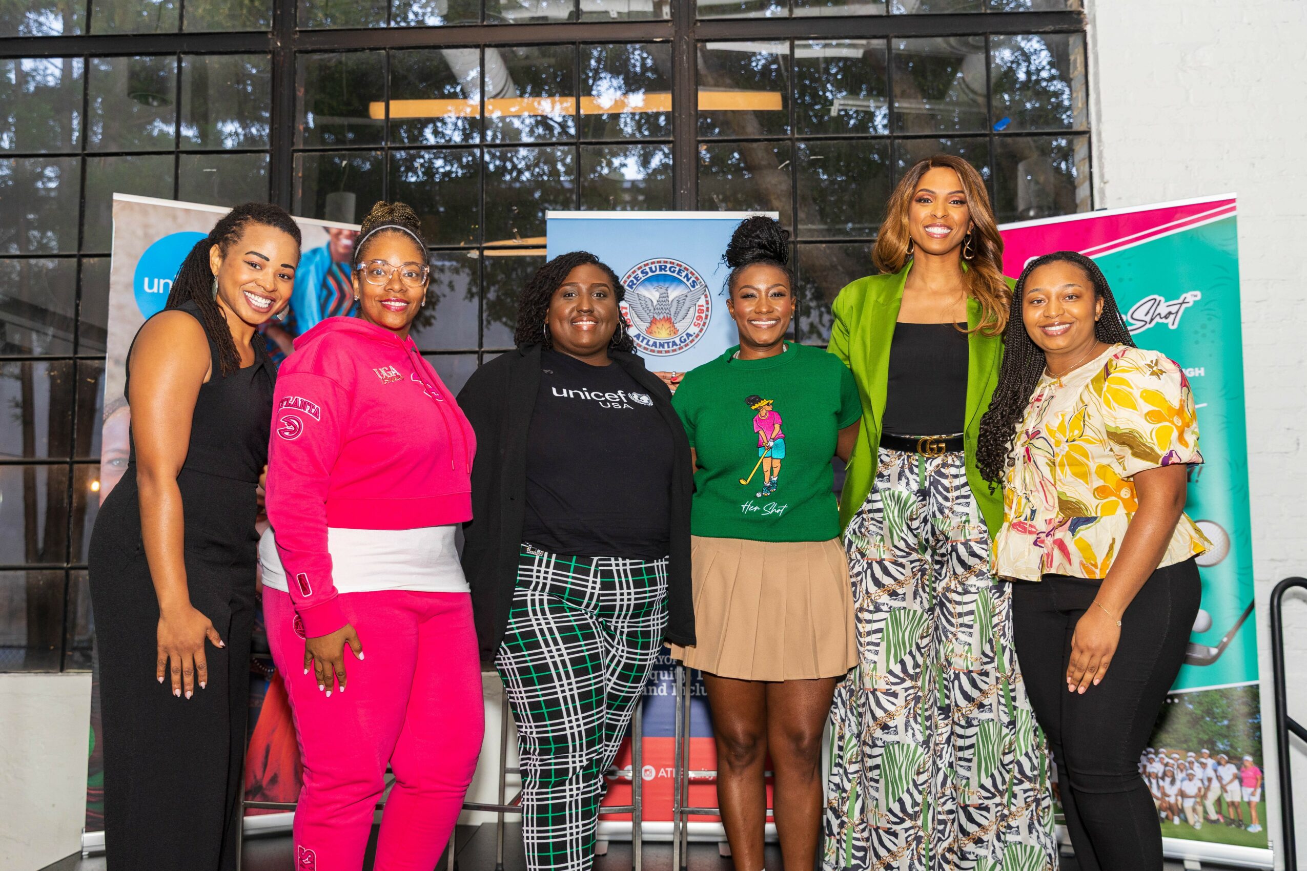 Six women standing together outdoors in front of a building with glass windows, smiling and posing for the photo.