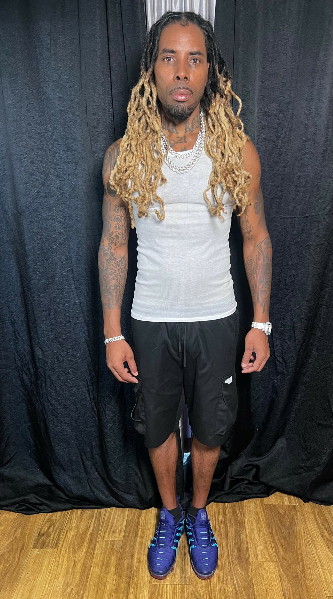 Man with long, blonde dreadlocks wearing a white tank top, black shorts, and blue shoes, standing against black curtains.