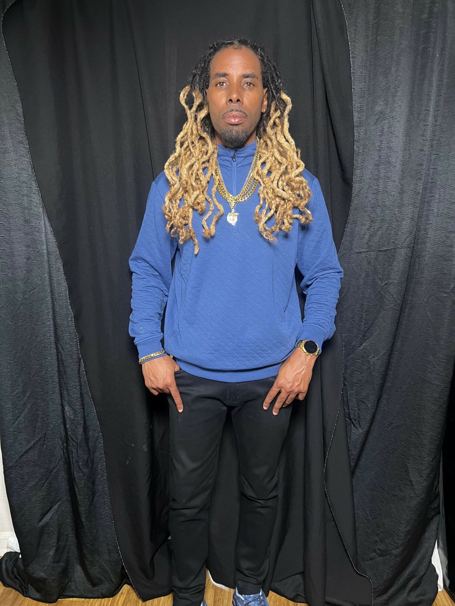Man with long dreadlocks wearing a blue sweatshirt and black pants, standing in front of black curtains.