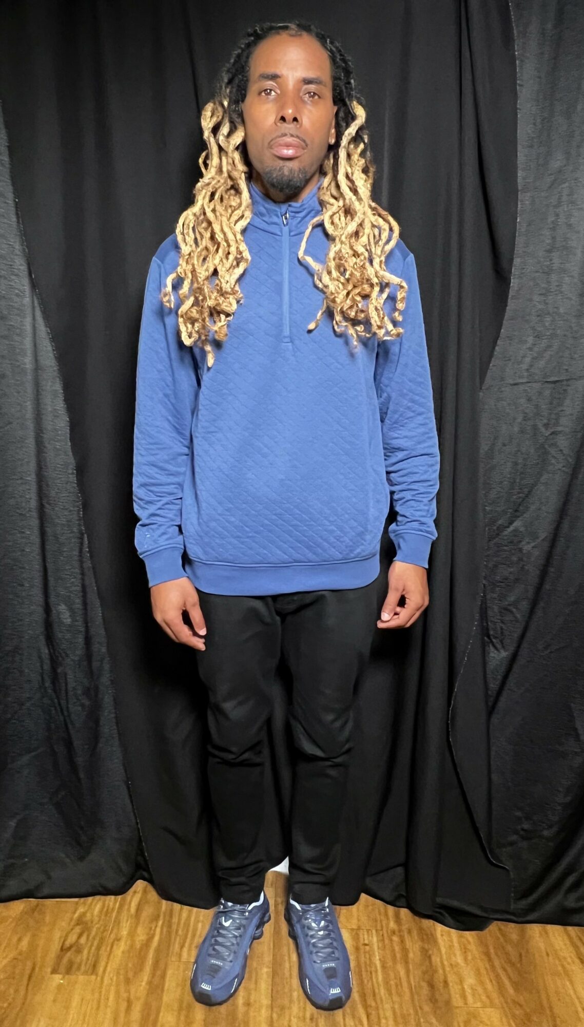 Young man with long, curly blond hair, wearing a blue sweatshirt, black pants, and blue sneakers, standing in front of black curtains.