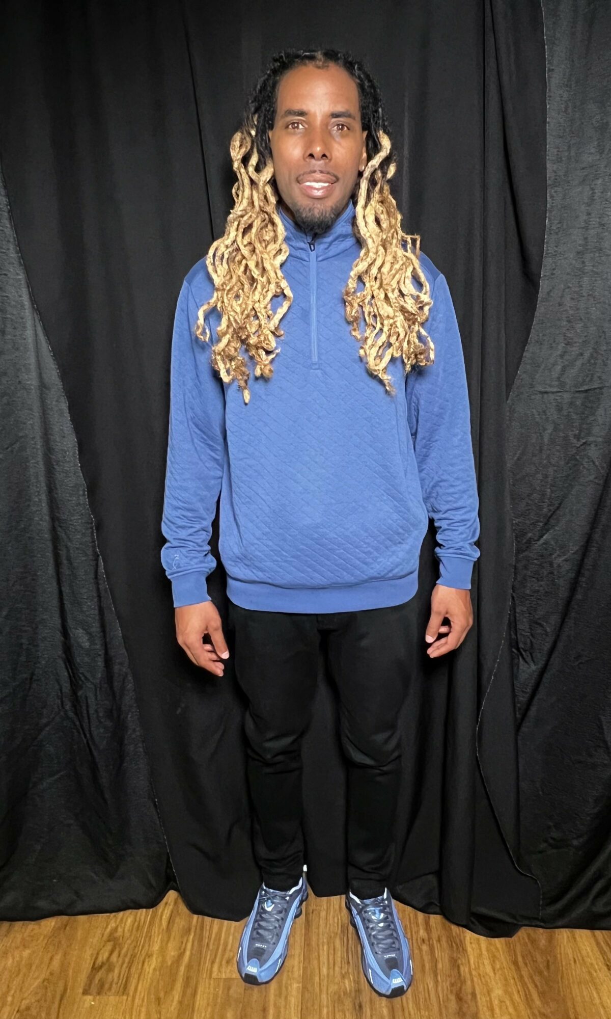 Person with long, curly blond hair wearing a blue sweatshirt, black pants, and blue shoes, standing on wooden floor against black curtains.