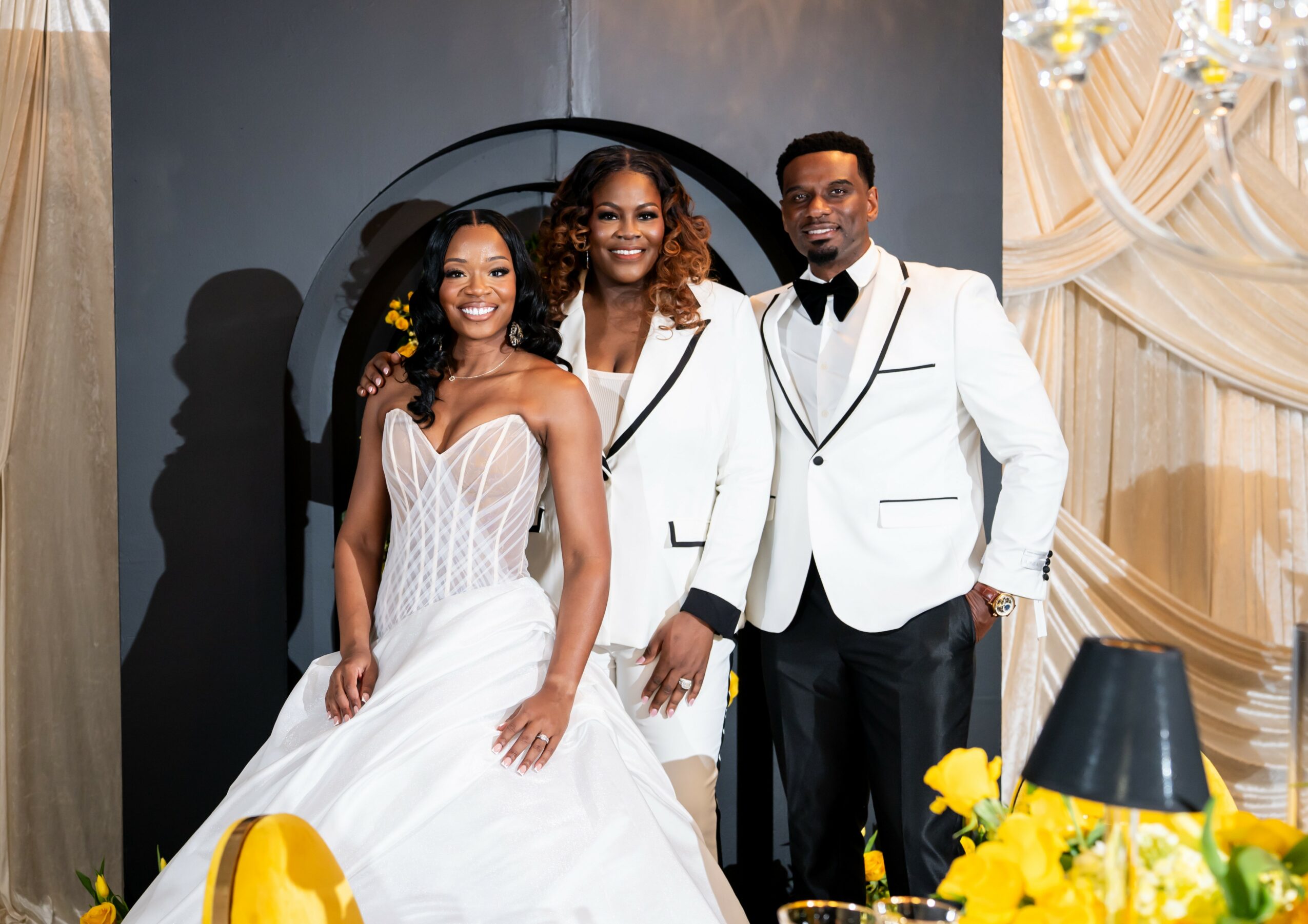 Three people dressed in formal attire, smiling, standing indoors with elegant decorations and yellow flowers.