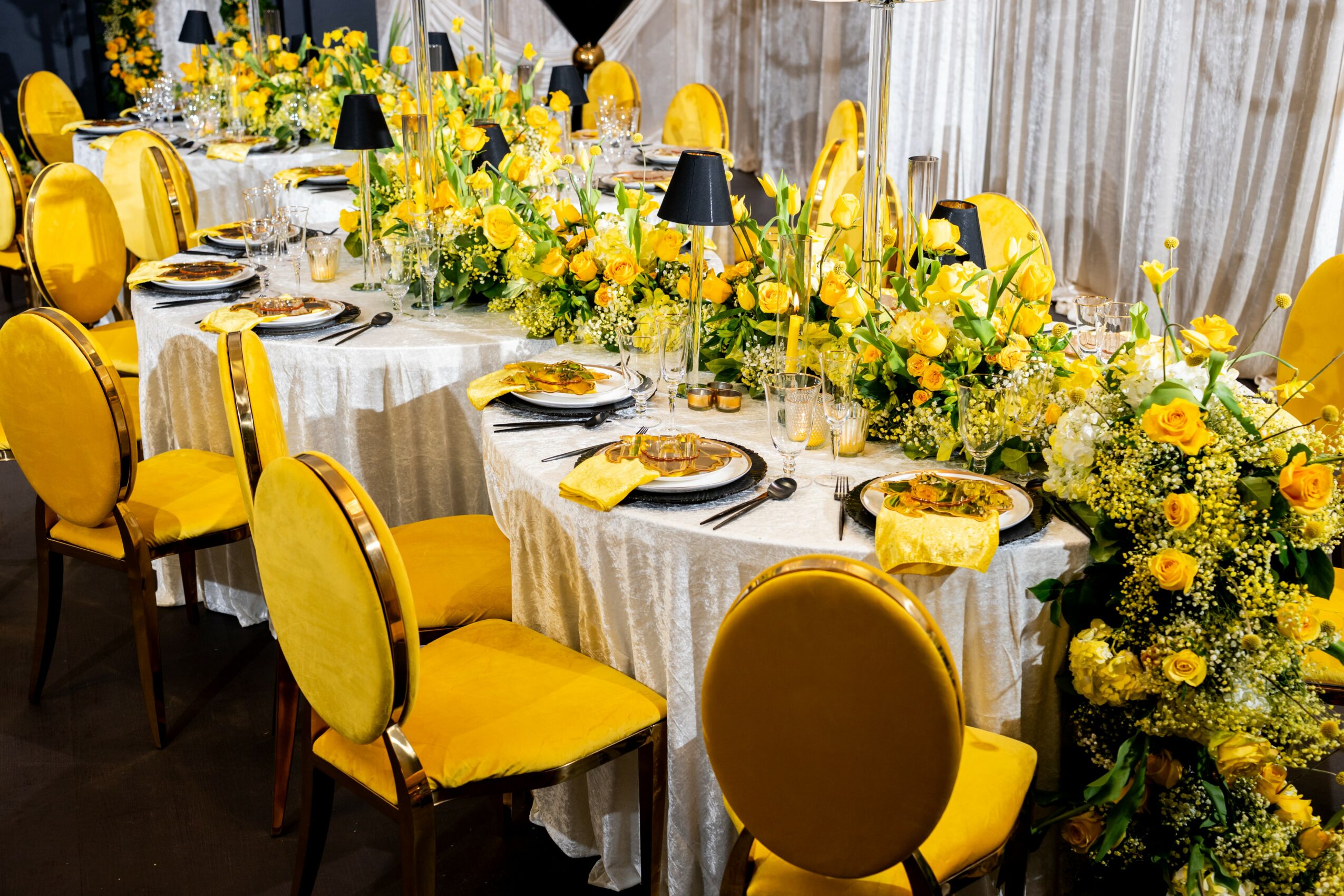 Long banquet table decorated with yellow flowers and yellow chairs, set with plates, glasses, and cutlery.
